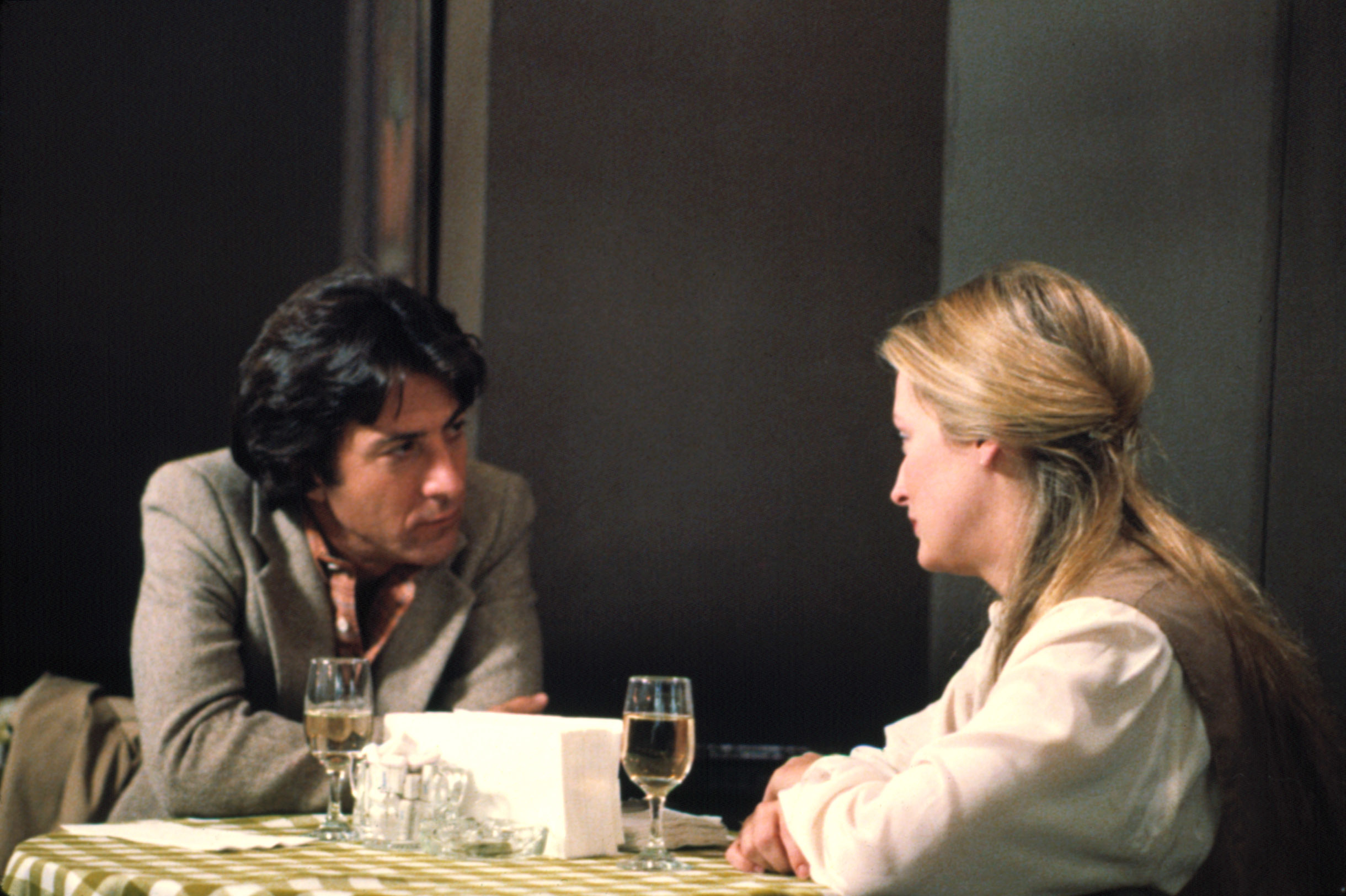 Dustin Hoffman and Meryl Streep are seated at a restaurant table, engaged in a serious conversation with glasses on the table