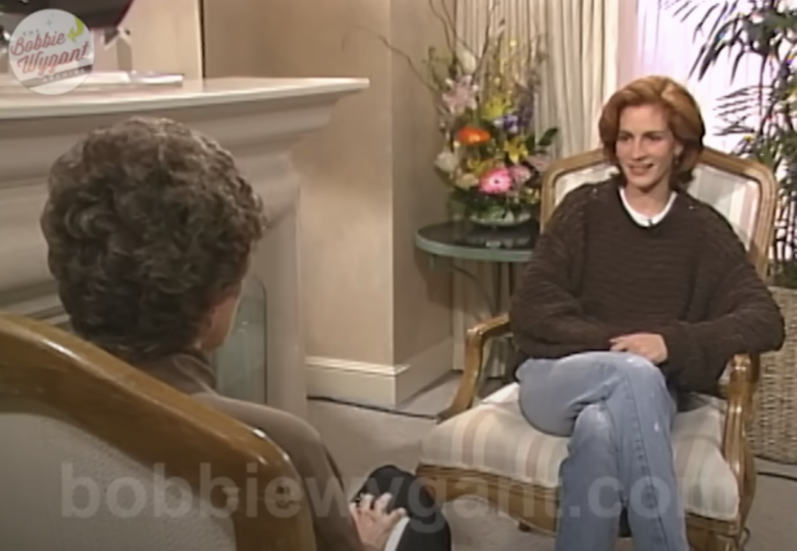 Julia Roberts, wearing a casual sweater and jeans, is being interviewed by Bobbie Wygant in a cozy setting with floral decor