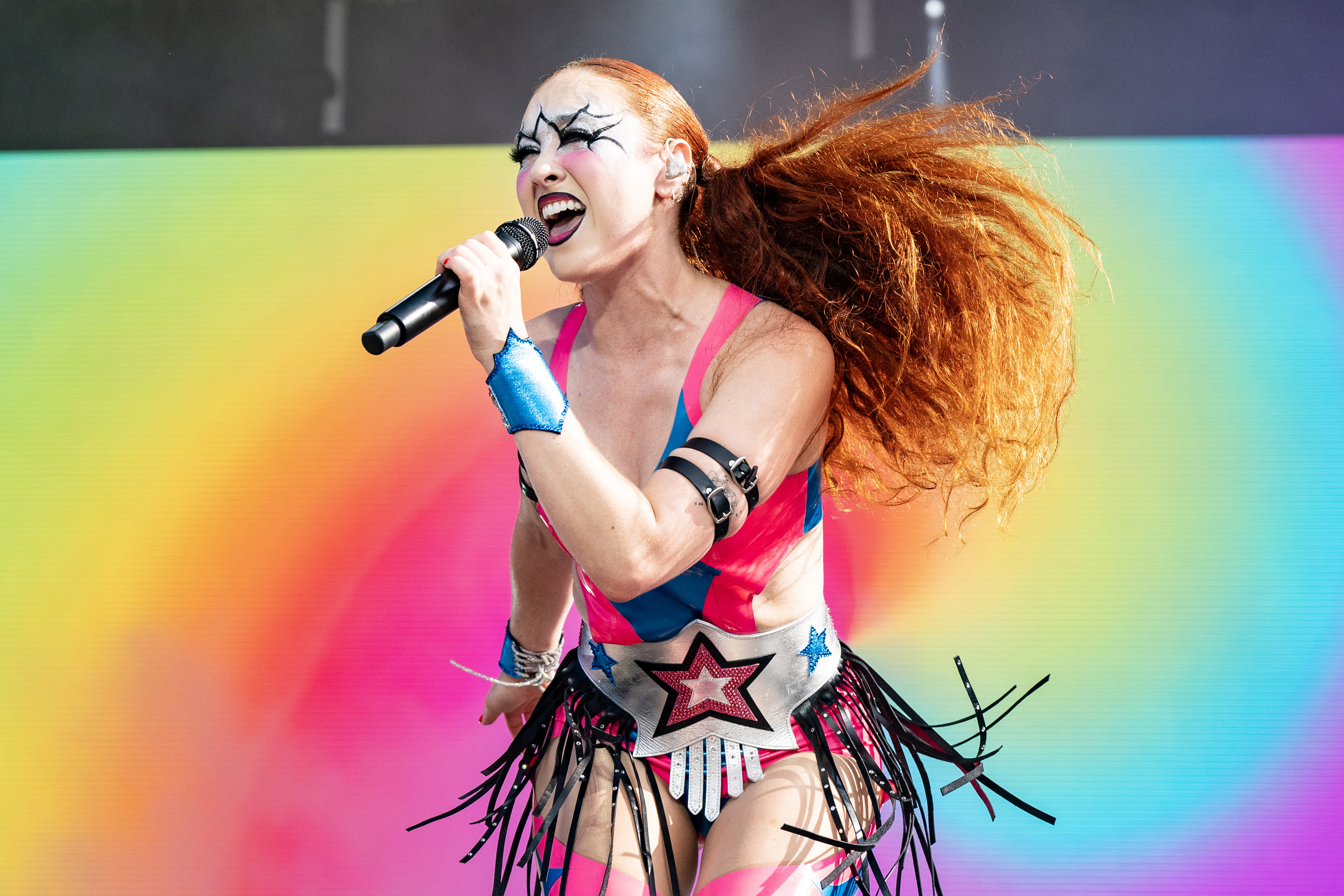 Chappell Roan with star face paint and red hair performs energetically on stage. She wears a vibrant costume with stars and fringes, holding a microphone