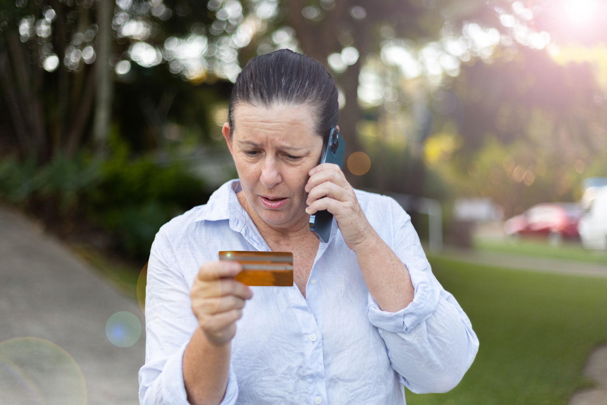 A woman in a white shirt holds a credit card in one hand and talks on a cellphone with a concerned expression