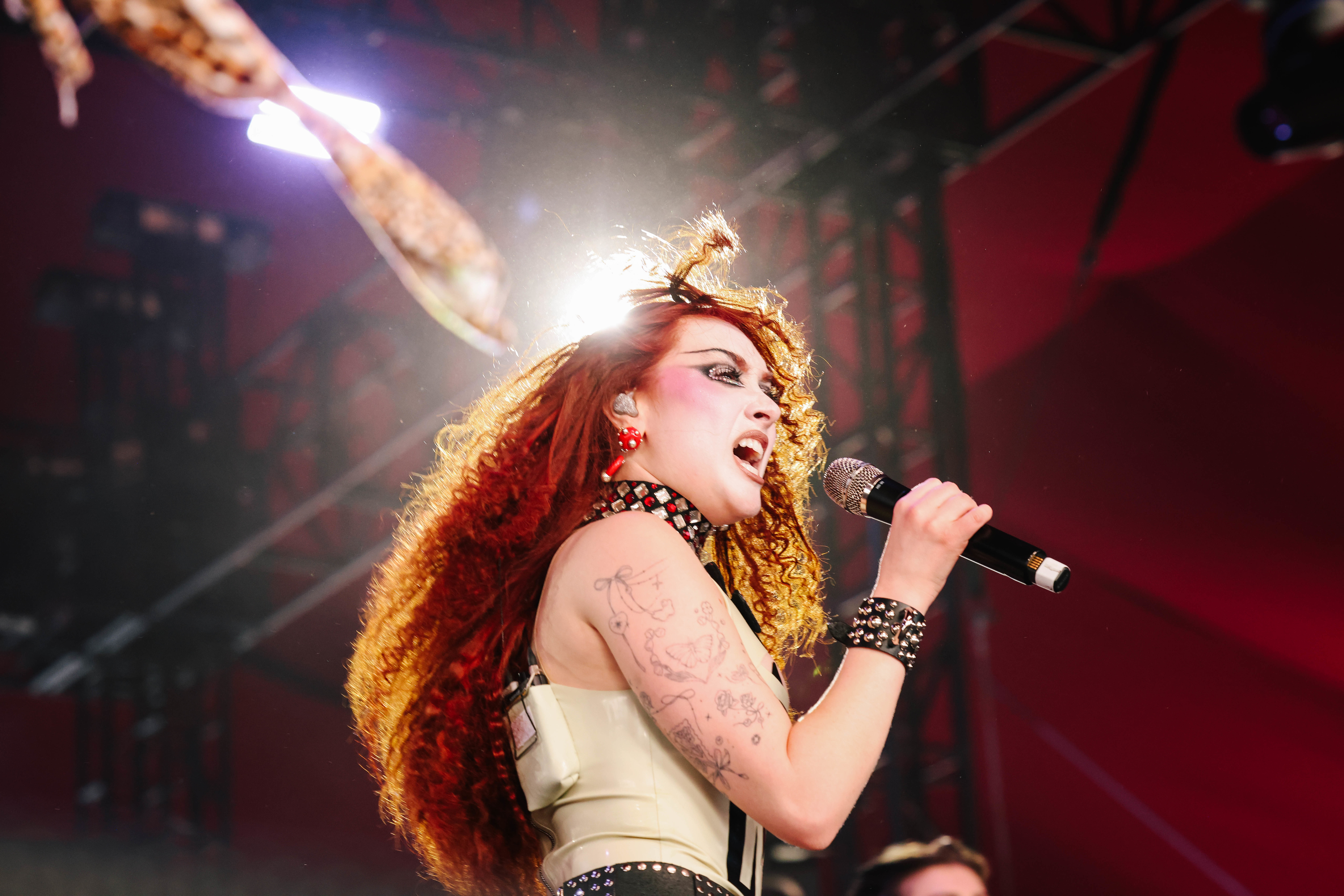 Chappell Roan with long curly hair and tattoos performs passionately on stage holding a microphone, wearing a studded outfit