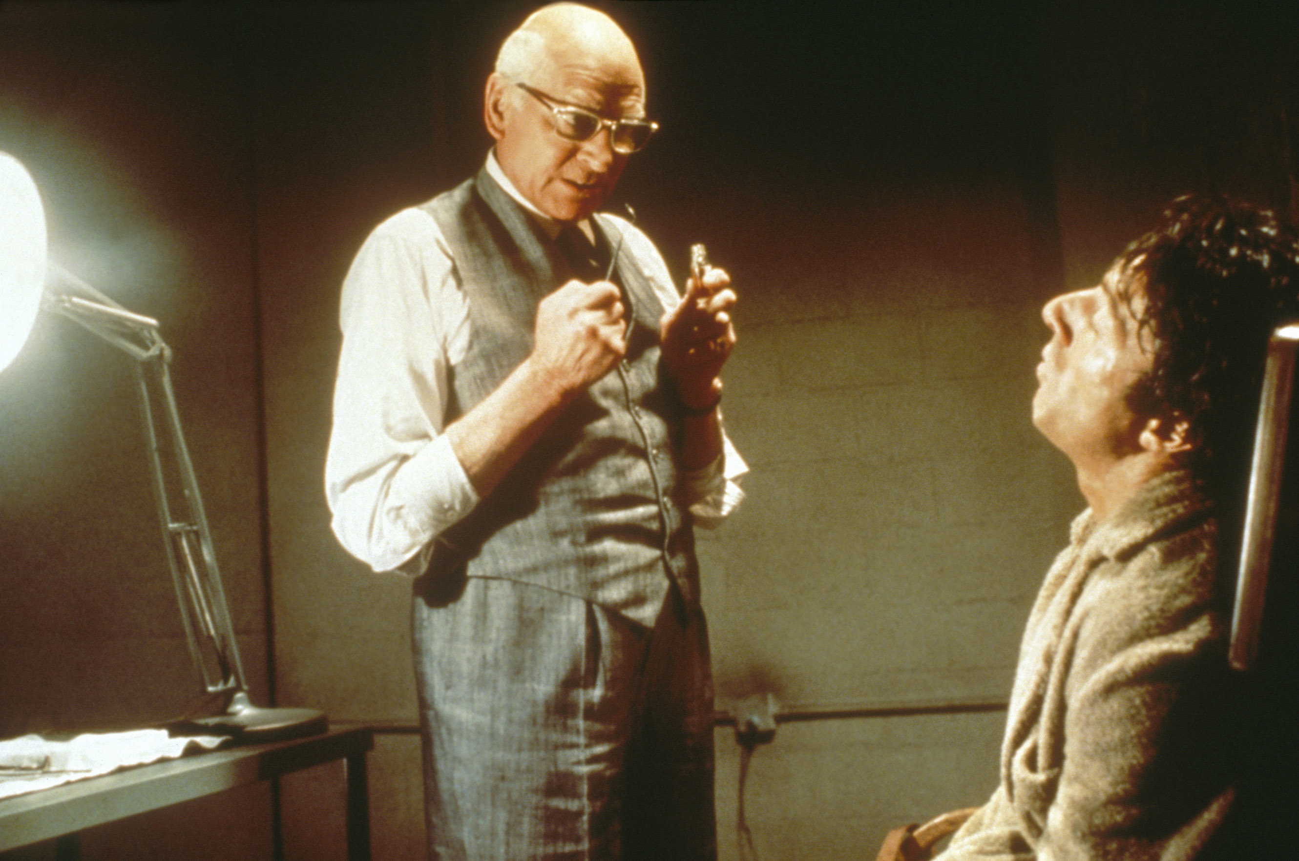 Lawrence Olivier, in a suit, holds tools standing over and facing Dustin Hoffman, who is seated in a chair in a dimly-lit room
