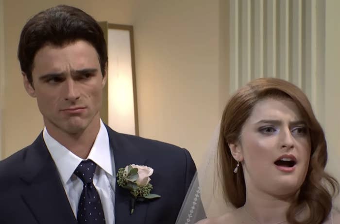 Characters from "Saturday Night Live" sketch in wedding attire, with a man in a suit and a woman in a bridal dress and veil expressing surprise