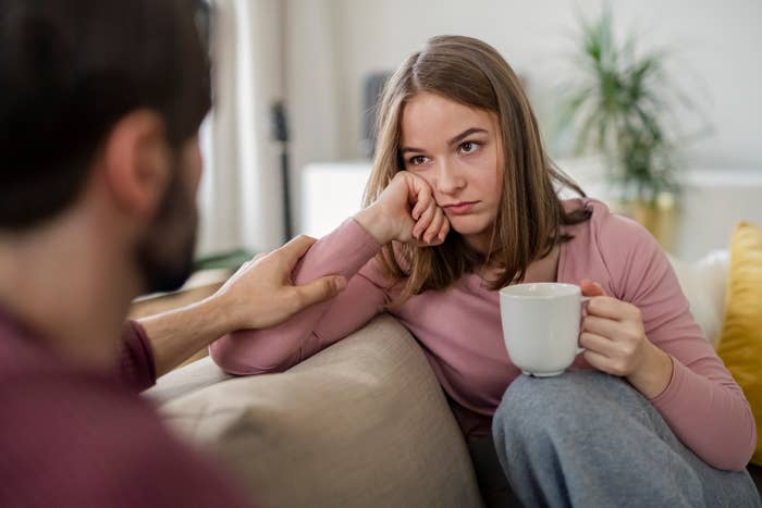 A woman holding a mug looks concerned, resting her head on her hand. A man sits beside her, gently holding her arm in a comforting gesture