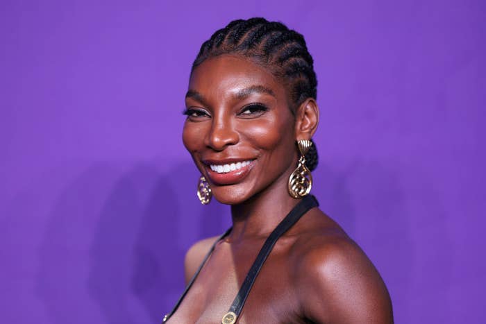 Michaela Coel is smiling at the camera while wearing braided hair and gold hoop earrings against a purple background
