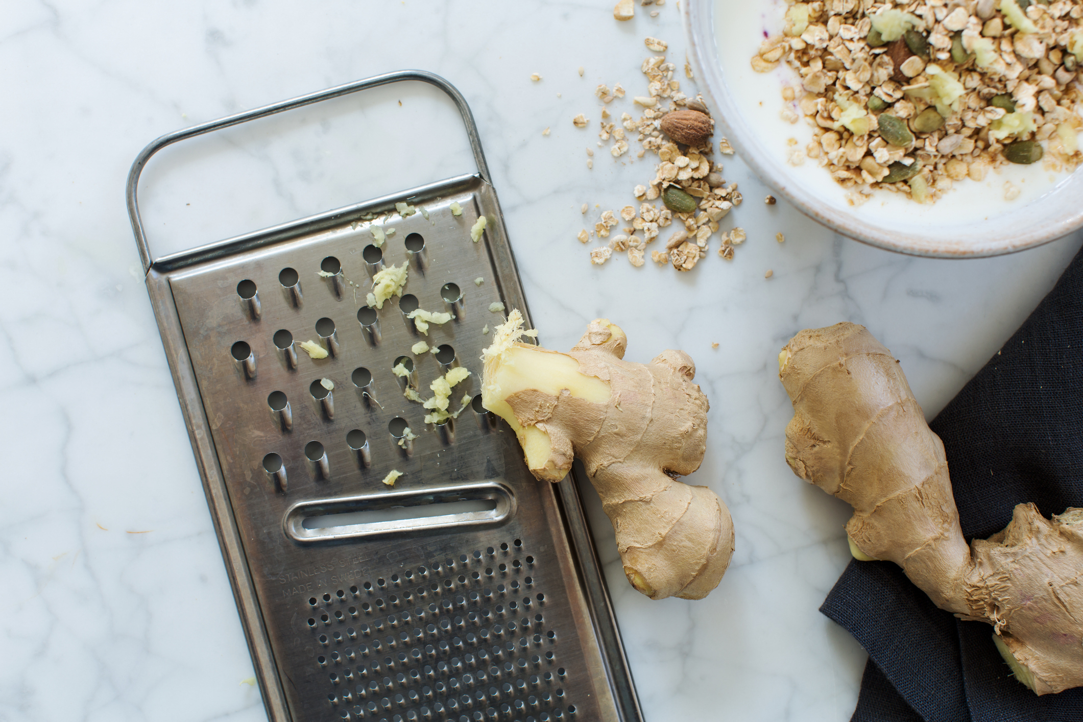 A metal grater with ginger pieces next to it, a bowl of granola and yogurt, and more ginger roots on a marble surface