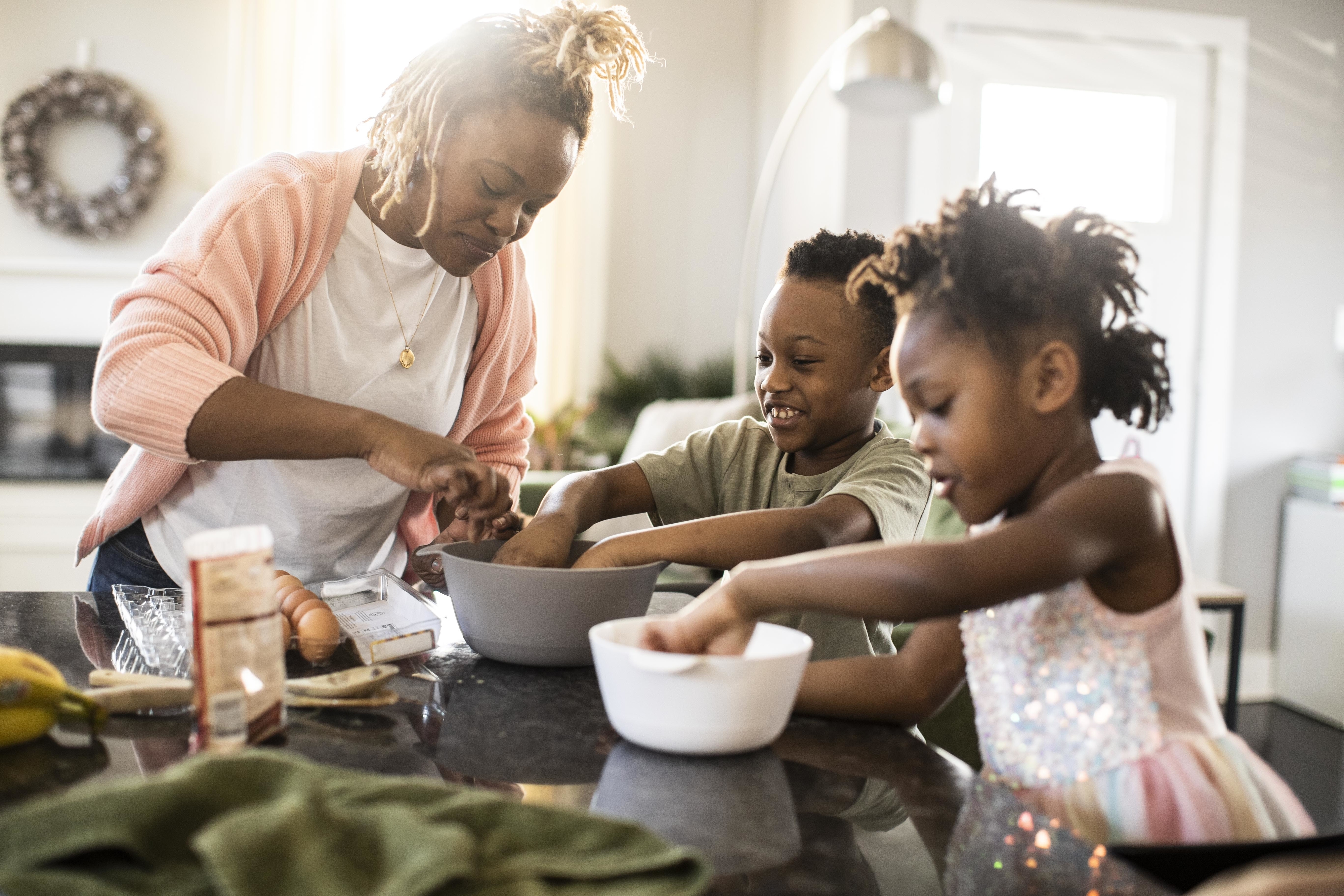 Woman with two children baking together in a kitchen. Various baking ingredients are on the counter, and all three are smiling and engaged in the activity