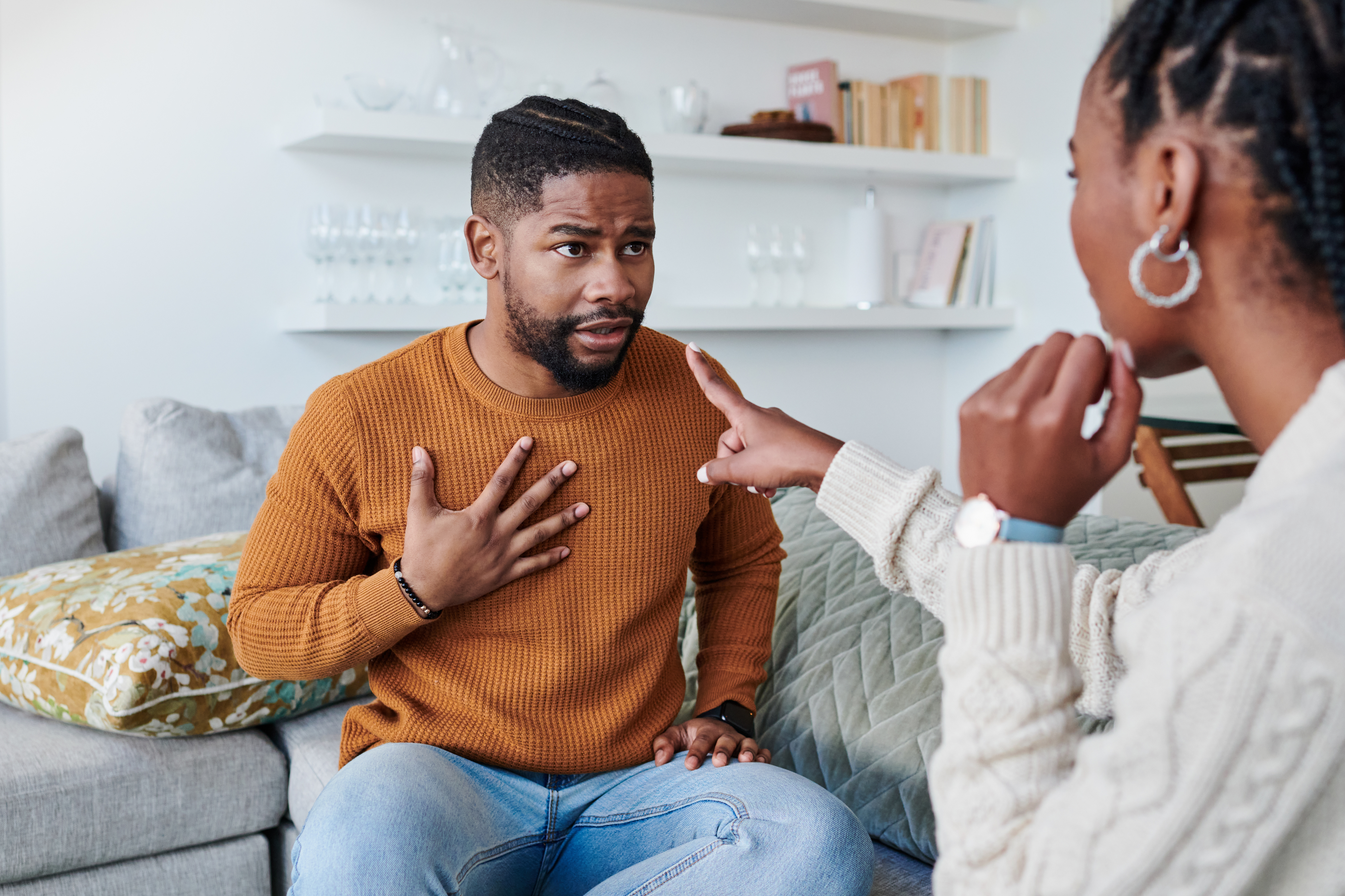 Man in a brown sweater looks concerned while being pointed at by a woman in a white sweater, sitting on a couch at home