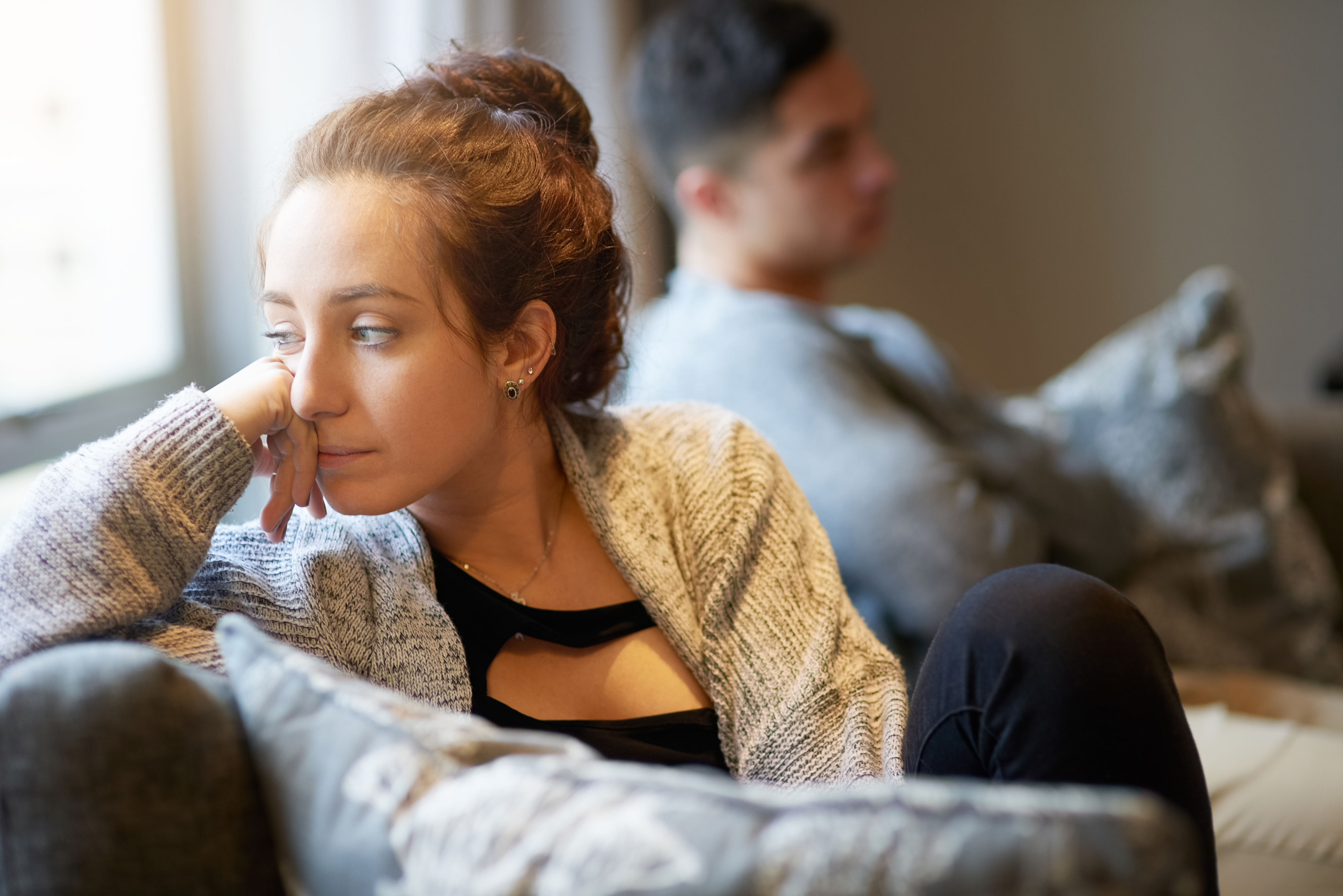 A woman with her hair in a bun looks out a window thoughtfully, while a man, sitting behind her on a couch, looks away in the opposite direction