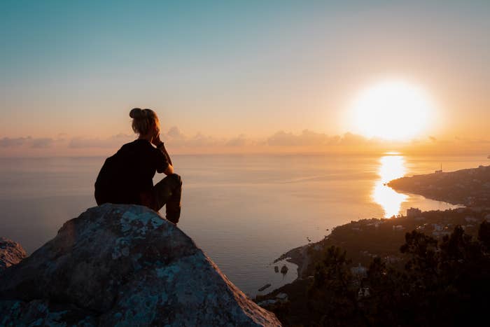 Person sits on a rock overlooking a scenic coastal view at sunset