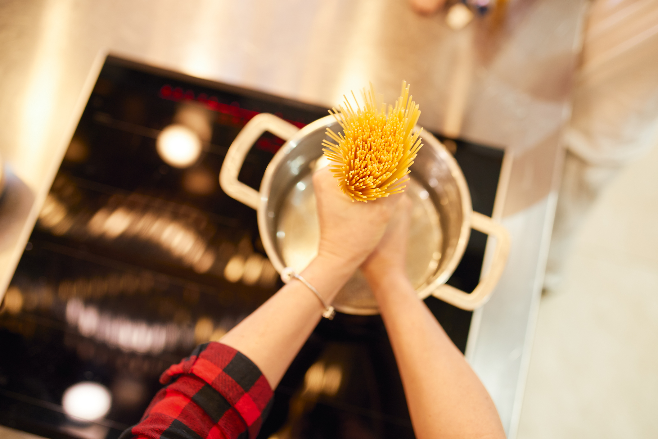 A person in a plaid shirt places uncooked spaghetti into a pot of boiling water on an induction cooktop