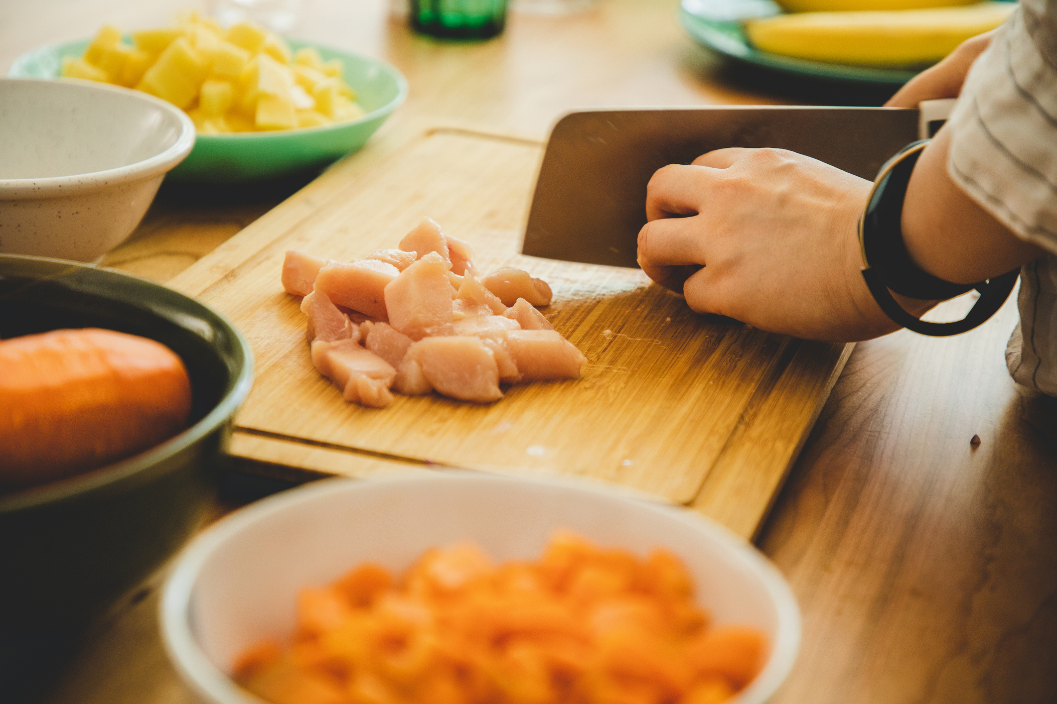 A person in a kitchen chops raw chicken on a wooden cutting board, surrounded by bowls of chopped vegetables