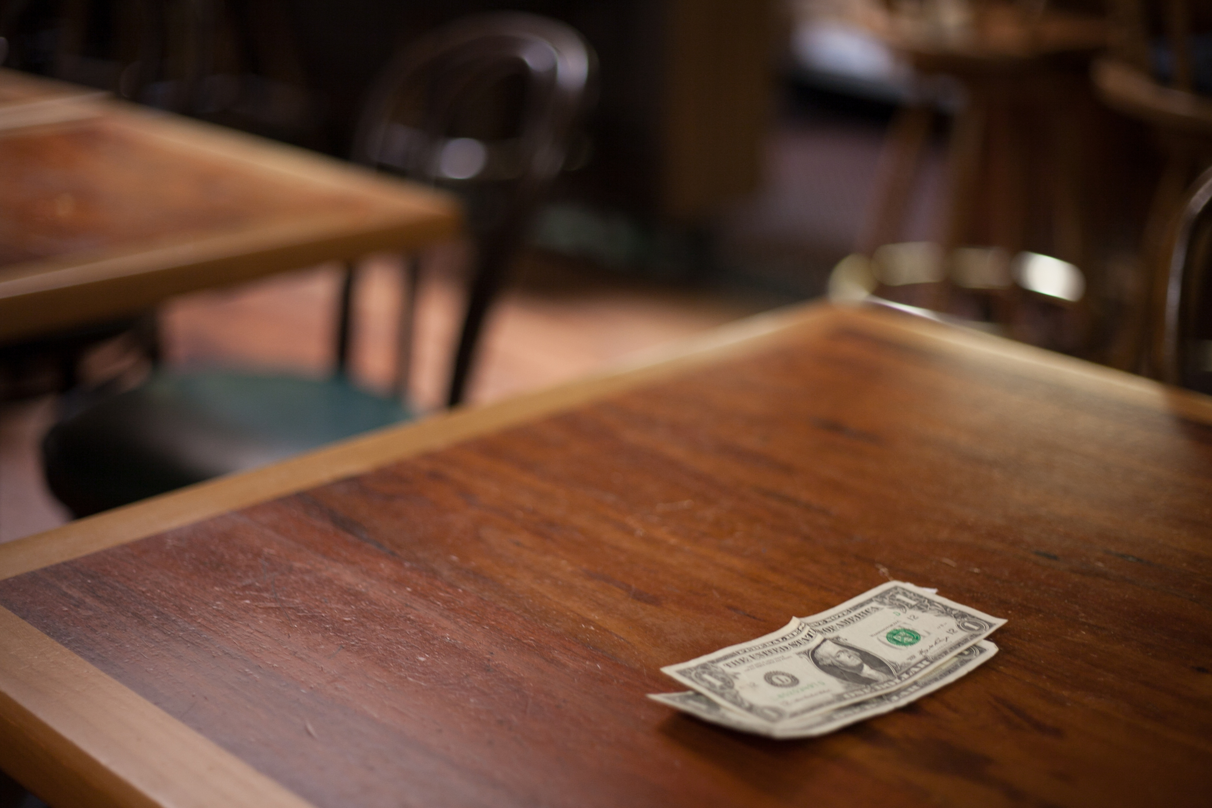 Two dollar bills are placed on a wooden table surrounded by empty chairs. No people are in the image