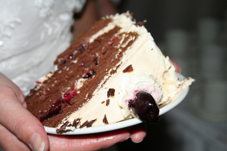 Close-up of a wedding cake slice held on a plate, featuring chocolate layers, cream frosting, and a cherry on top