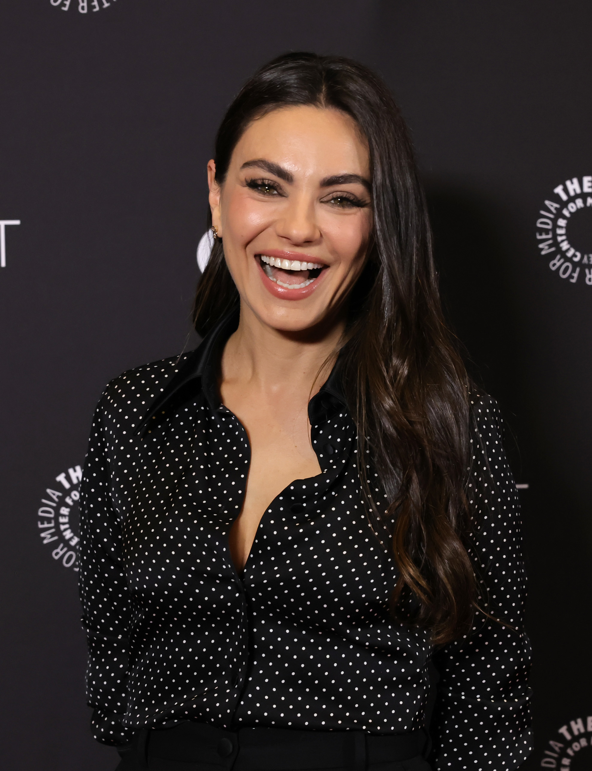 Mila Kunis, smiling, wears a black polka dot blouse at a celebrity event