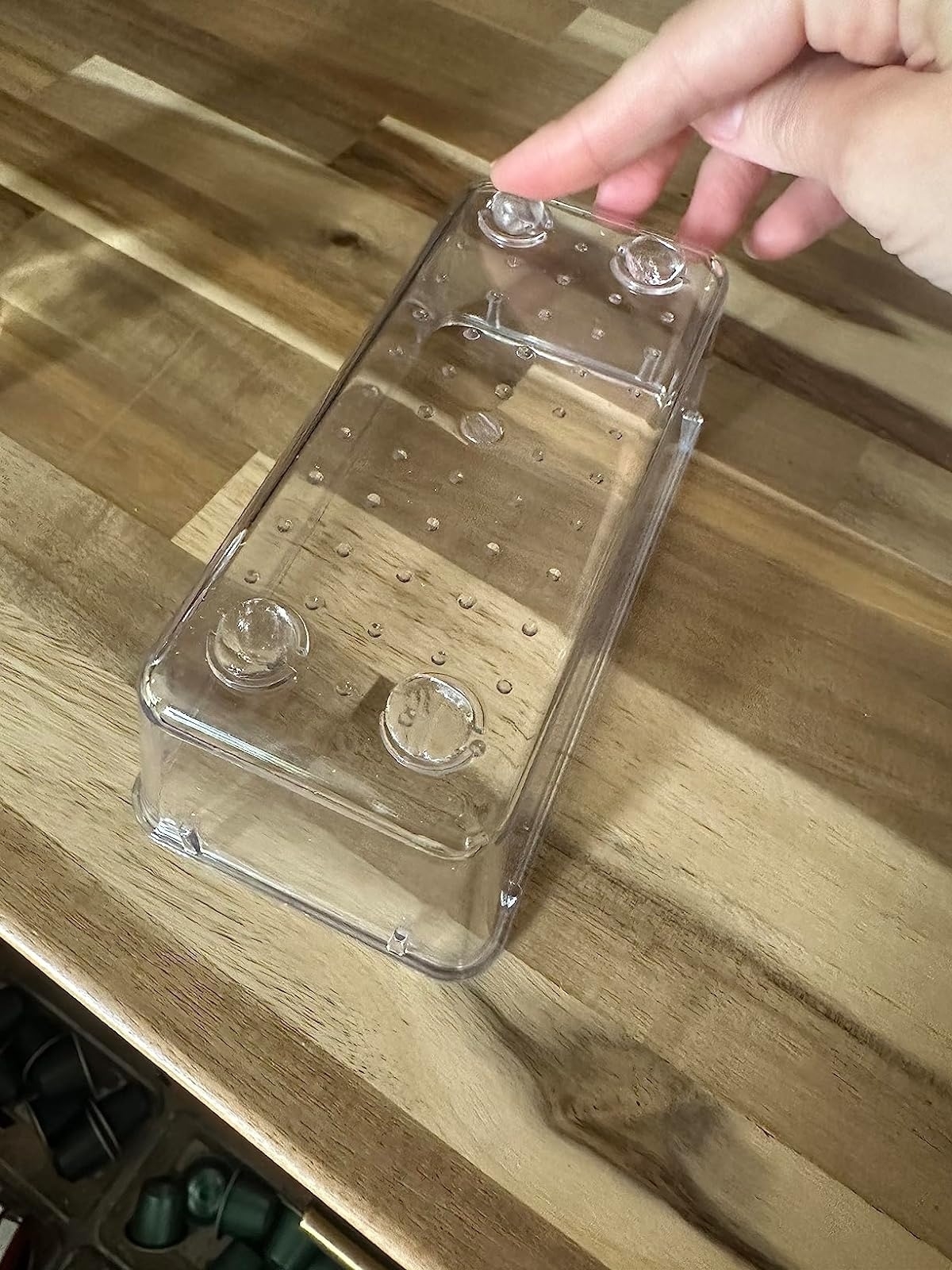 Hand holding a clear plastic container with suction cups, likely for kitchen or bathroom organization, placed on a wooden surface