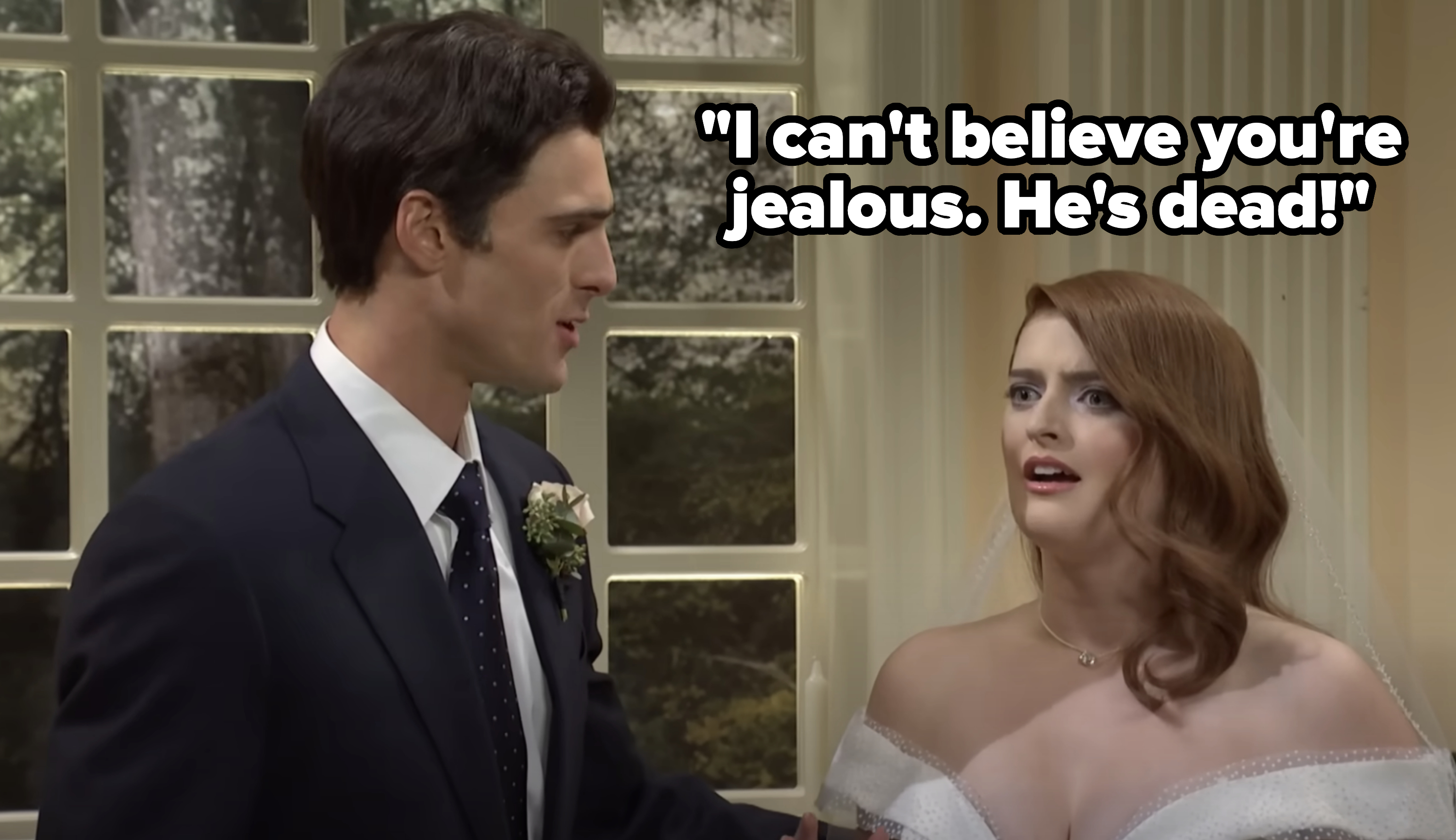 A bride in an off-the-shoulder wedding dress and a groom in a suit with a boutonniere are looking at each other inside a room with tall windows