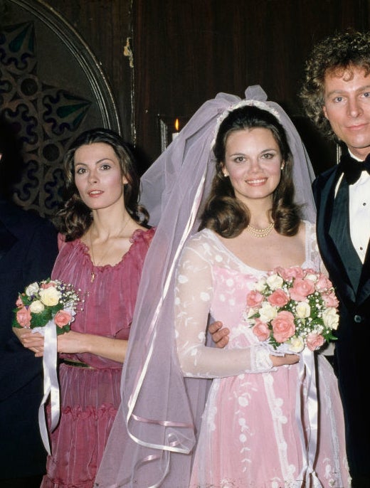 Bride wears long, pink dresses with veils and bouquets, standing next to a groom