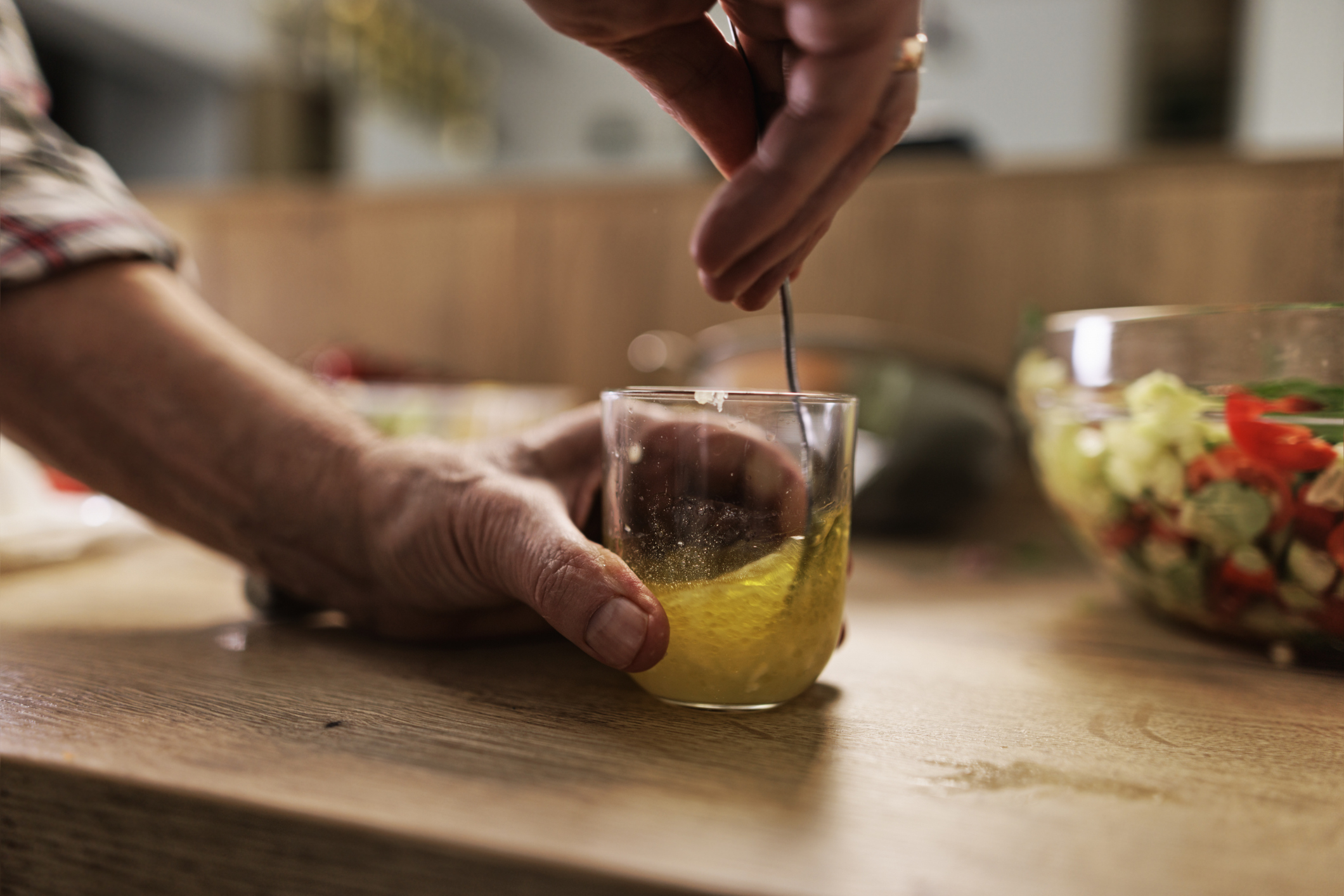 A person stirs a yellow liquid in a glass with a spoon. A bowl of chopped vegetables is in the background on the wooden countertop