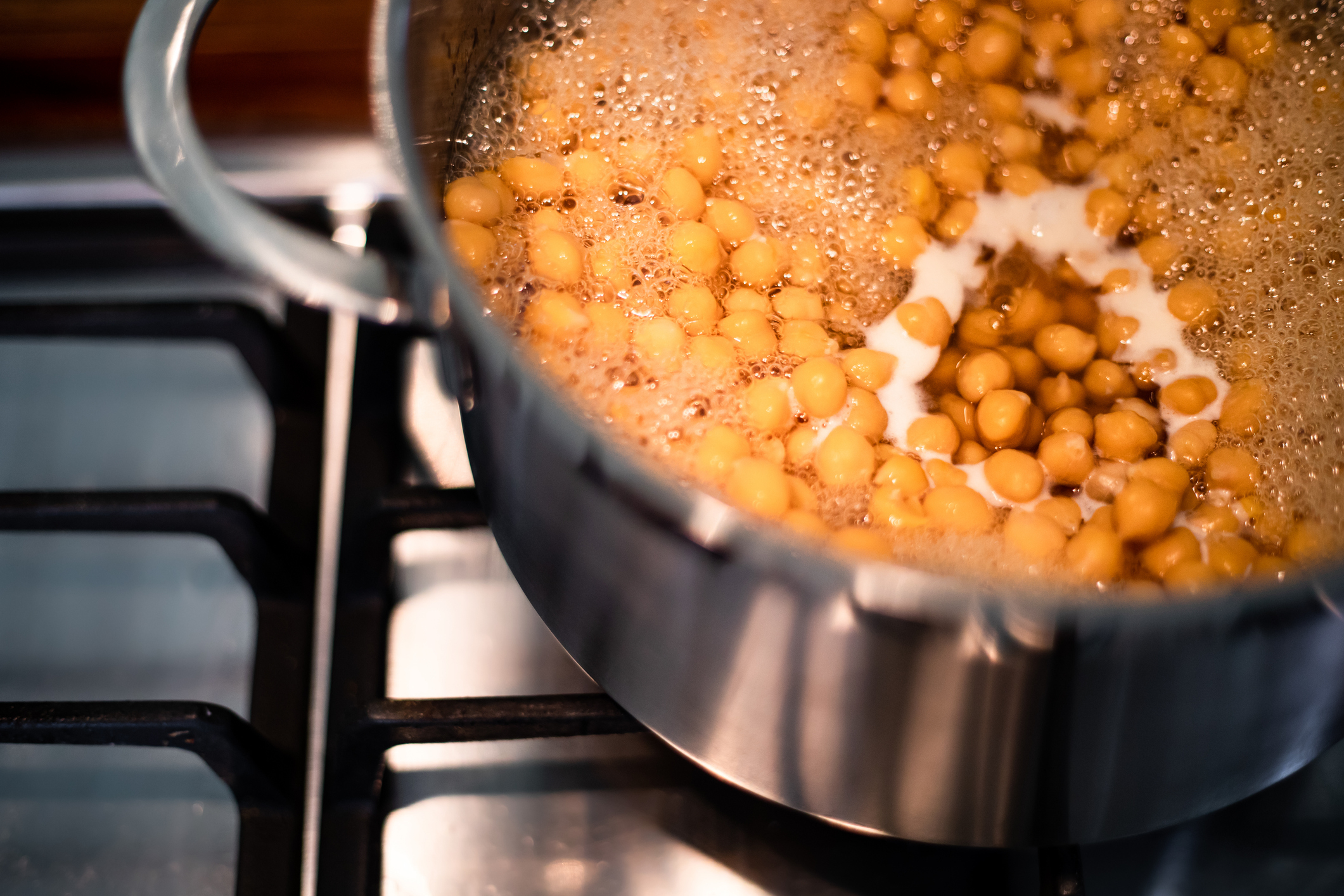 Chickpeas are boiling in a stainless steel pot on a stovetop, with steam and bubbles rising from the surface