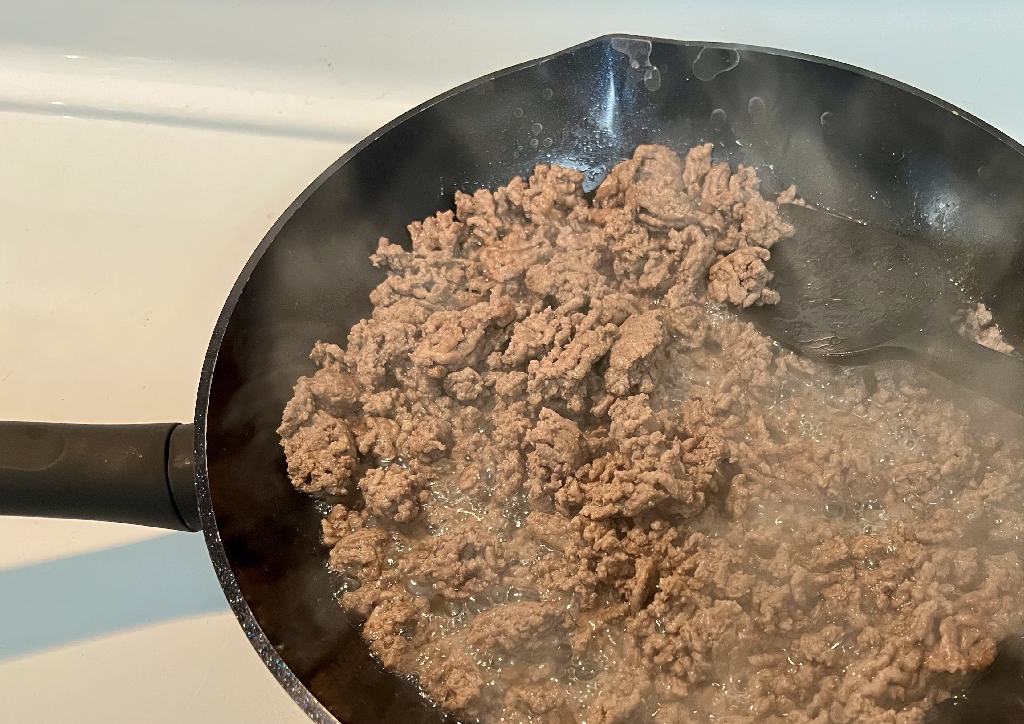 Ground beef cooking in a black frying pan with visible steam rising. A black spatula is partially seen on the right side