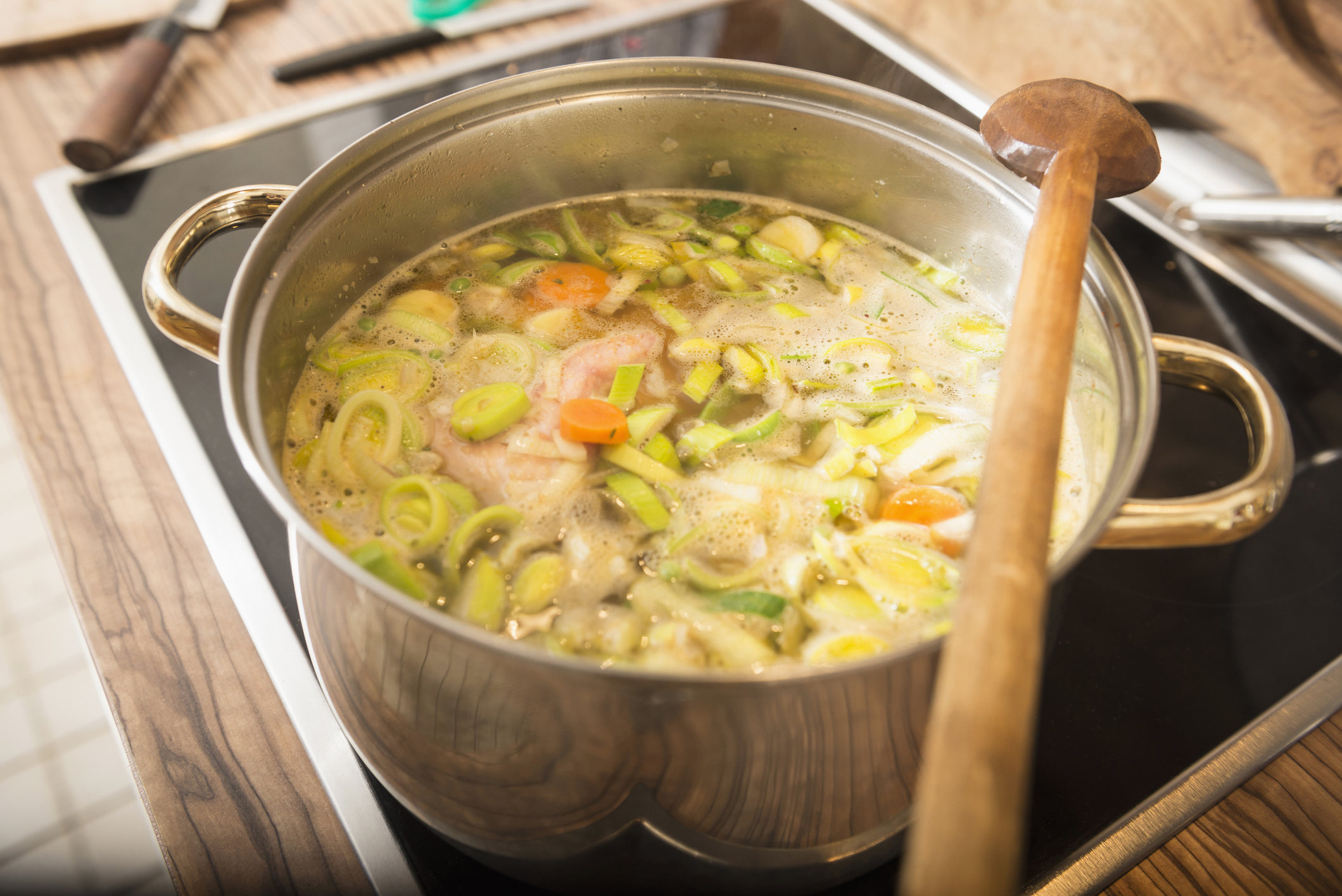 A pot on a stove filled with simmering soup that includes vegetables like carrots, leeks, and zucchini. A wooden spoon rests across the pot's handles