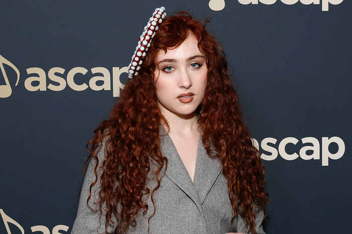 Woman with long curly hair and a headband stands in front of an ASCAP backdrop, wearing a gray blazer