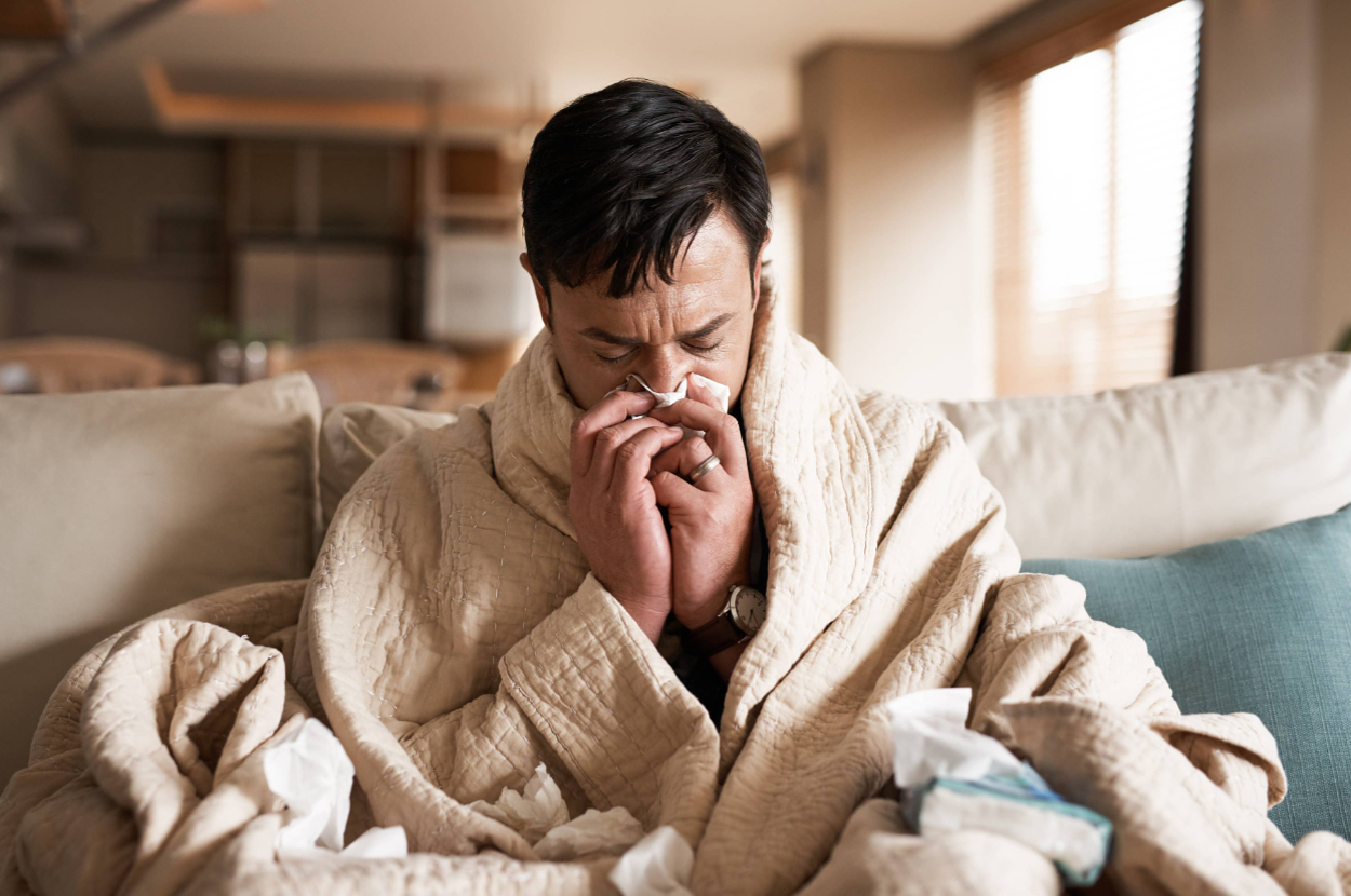 A person wrapped in a blanket is sitting on a couch, blowing their nose with tissues scattered around
