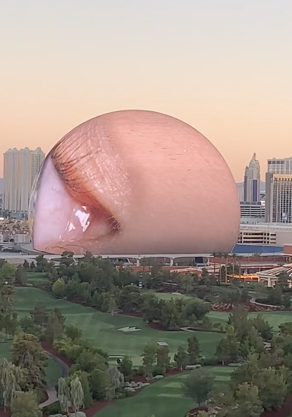 Close-up of a giant eye projected on a spherical screen, located among city buildings and greenery during sunset
