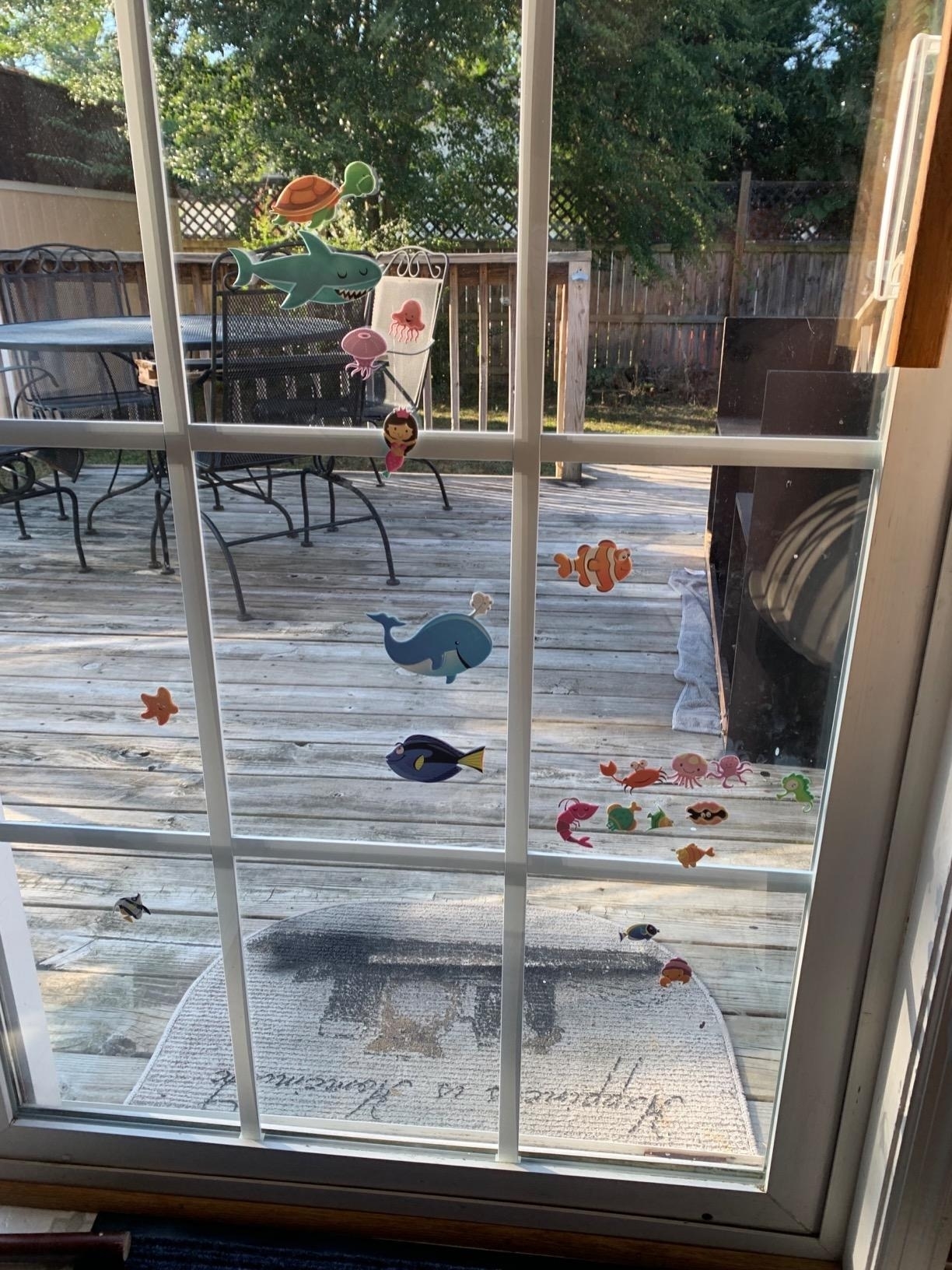 Outdoor deck viewed through a glass door, adorned with colorful sea creature stickers including fish, turtle, whale, and shells. Furniture and grill visible outside