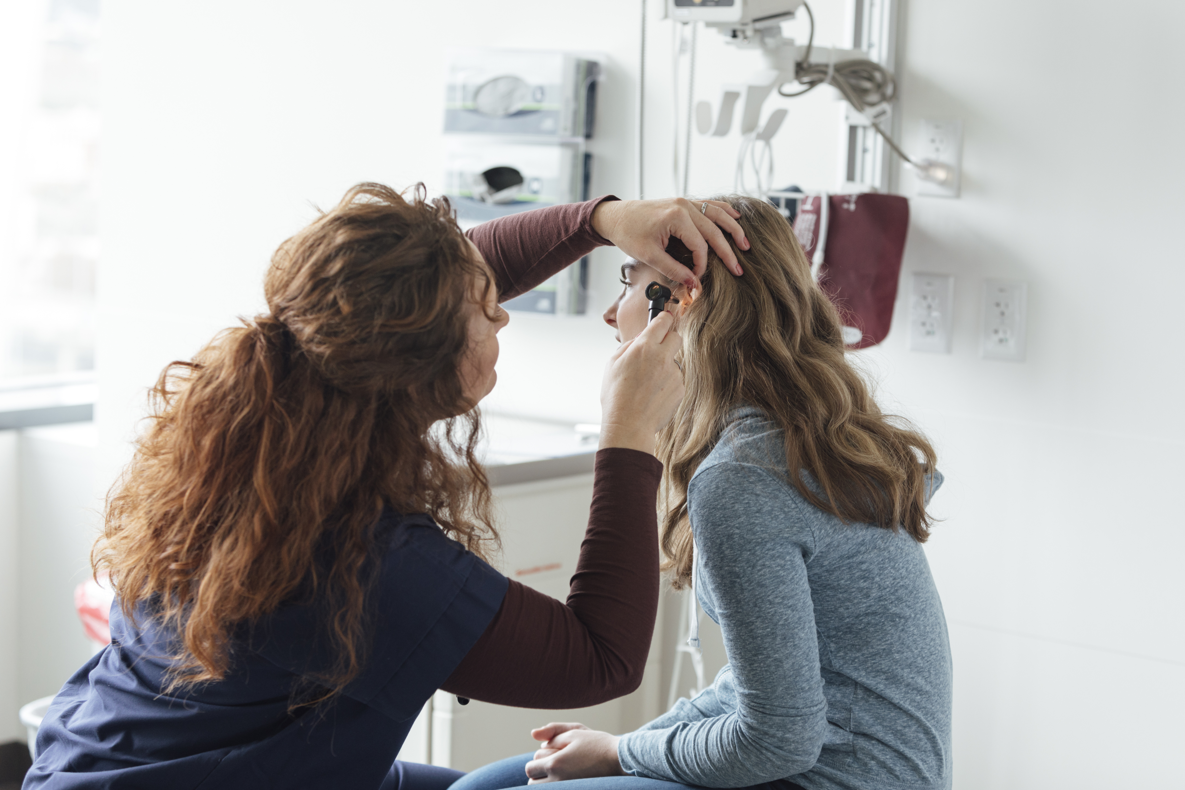 A healthcare professional examines a patient's ear in a clinic. Medical equipment is visible in the background