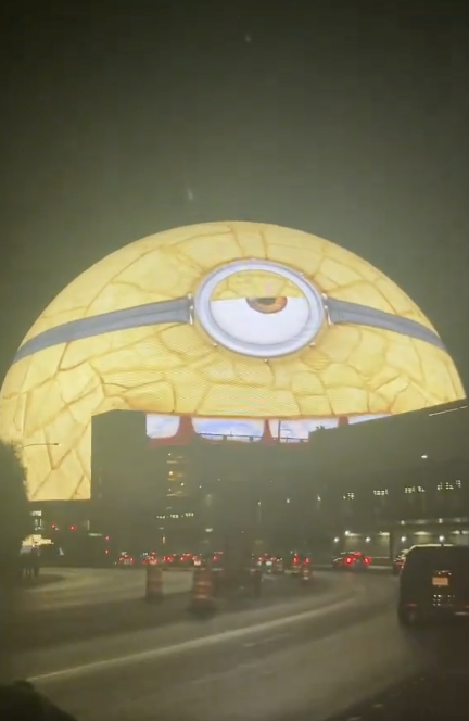 A large dome resembling a Minion from Despicable Me is illuminated at night, towering over a busy street with moving cars