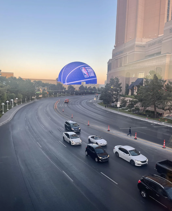 A wide street in a city with sparse traffic. A giant digital billboard in the distance displays an NBA Summer League design. People walk on the sidewalk