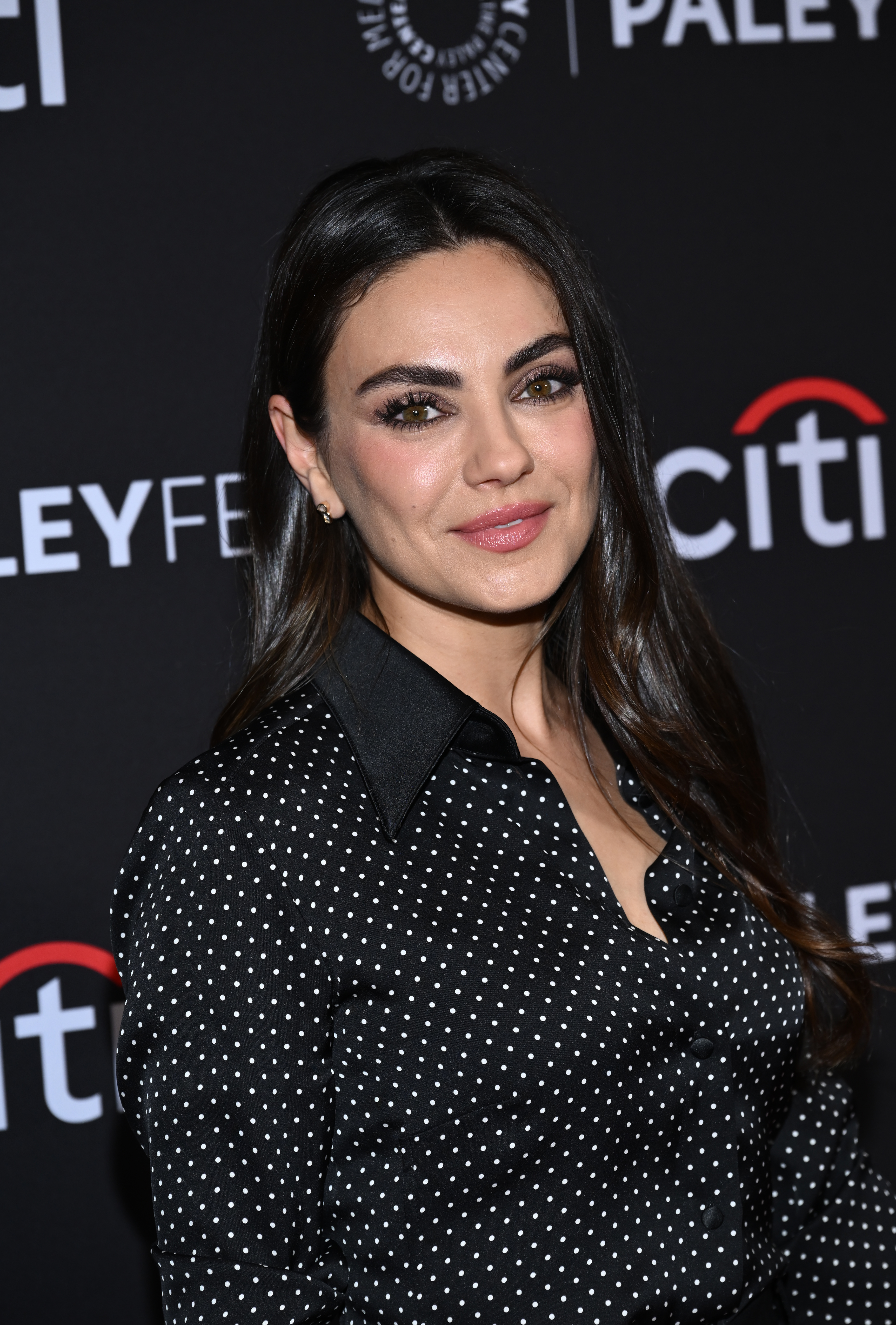 Mila Kunis at a PaleyFest event, wearing a black blouse with white polka dots