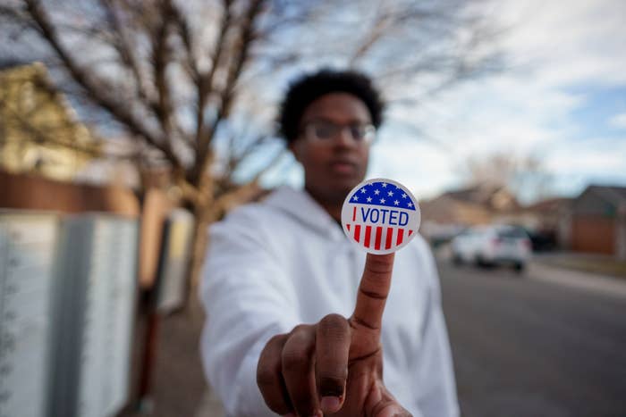 A person wearing glasses and a white hoodie holds up an &quot;I Voted&quot; sticker on their finger