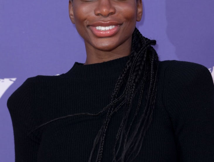 Michaela Coel smiles at the camera while posing on the red carpet in a stylish black outfit with braided hair