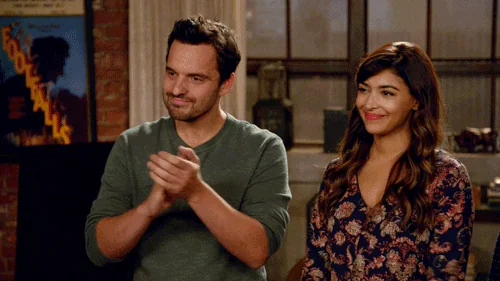Jake Johnson and Hannah Simone are seen standing and clapping, both smiling warmly in a scene from the show New Girl