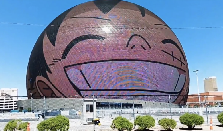A large spherical building displays a smiling, animated face on its exterior screen, seen from a distance with some greenery and structures in the foreground