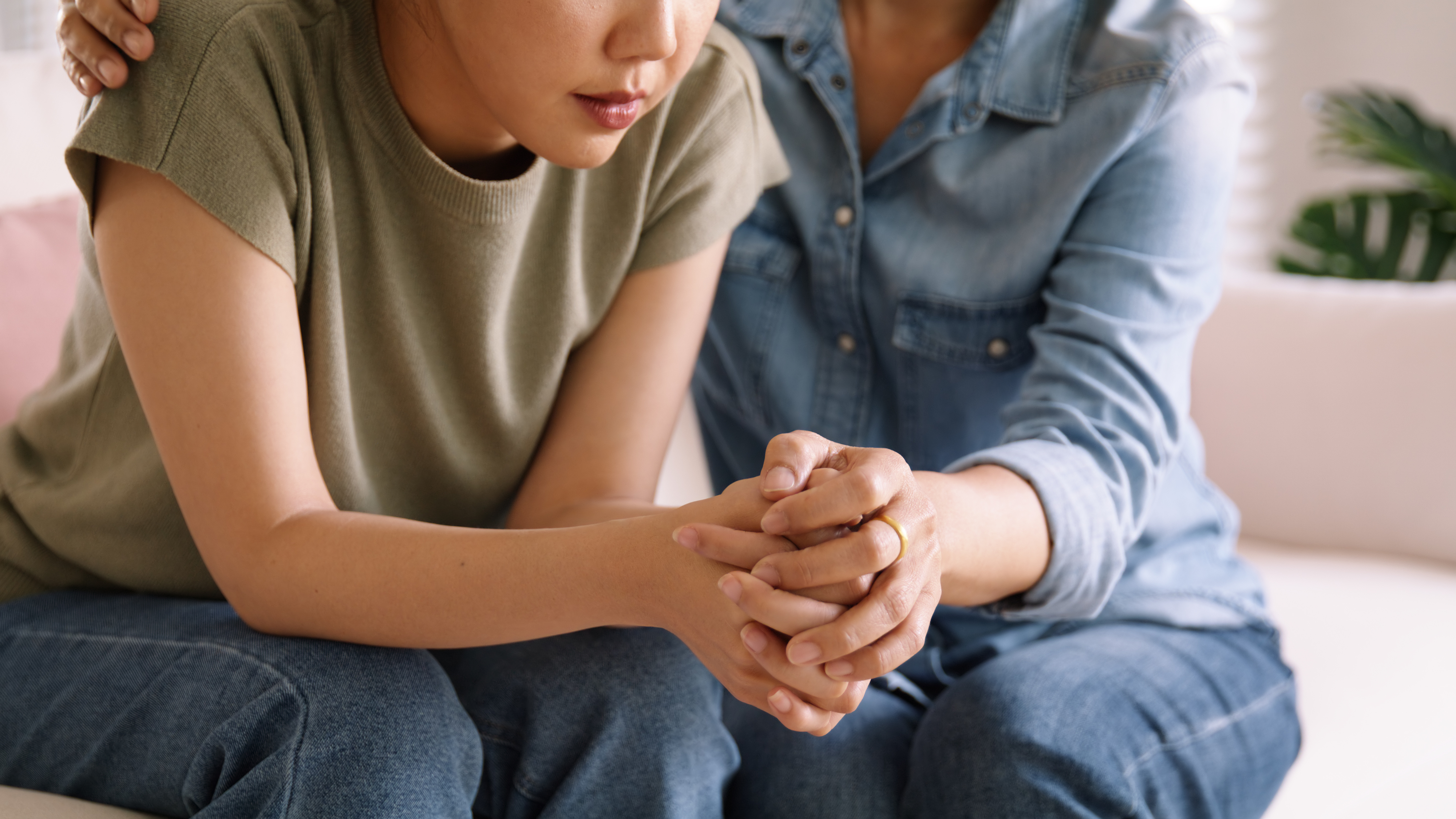 Two people sit closely on a couch, with one comforting the other by holding their hands. Their faces are not fully visible