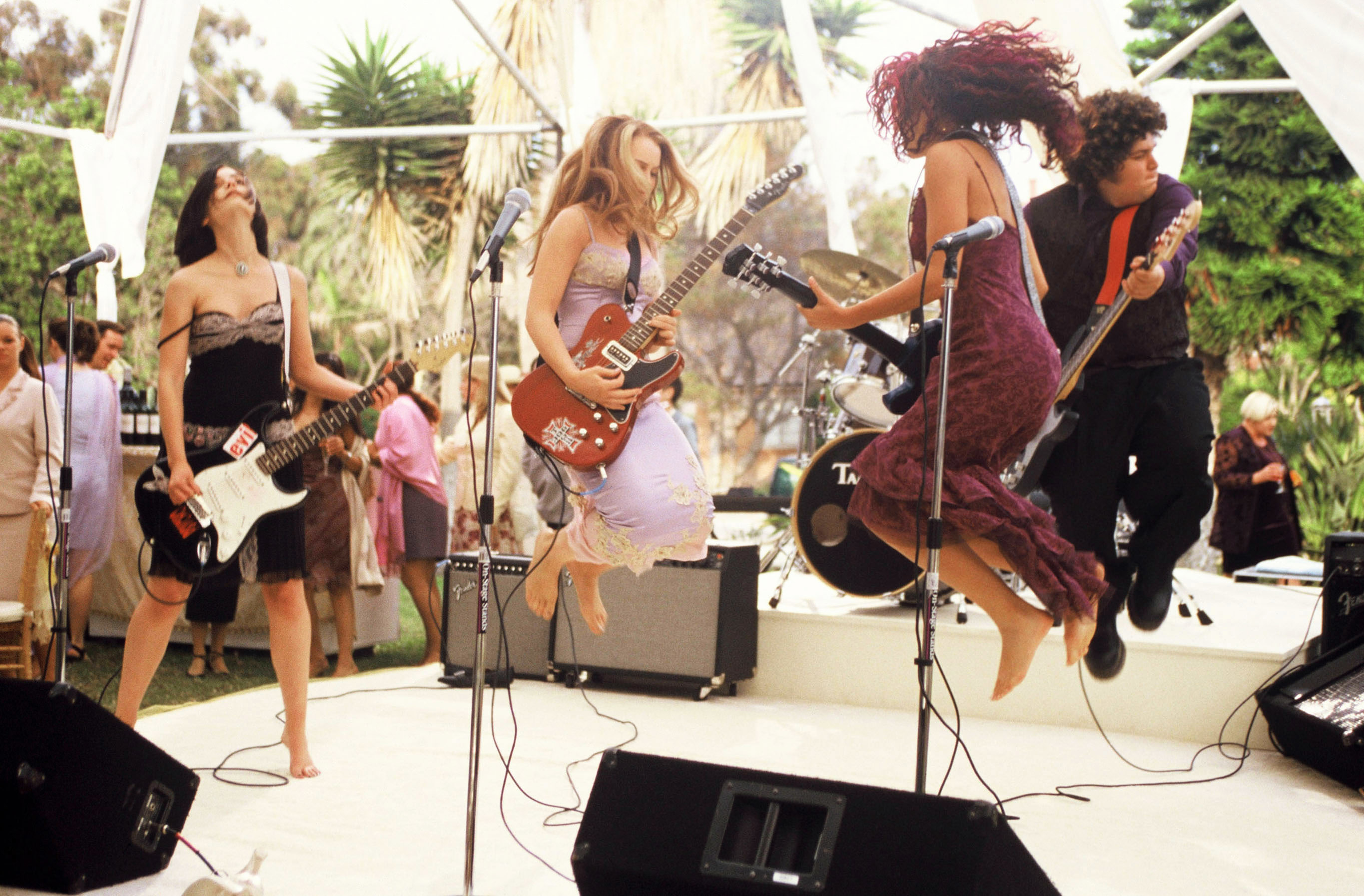 Liv Tyler, Jennifer Connelly, Coyote Shivers, and Gina Gershon perform energetically in a band, all jumping mid-air on stage with instruments