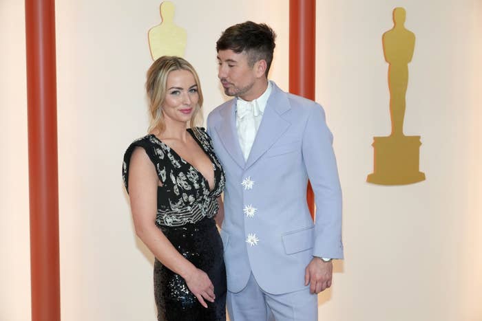 Alyson Kierans in a patterned dress and Barry Keoghan in a light-colored suit with floral details, pose together at an event with Oscar statues in the background