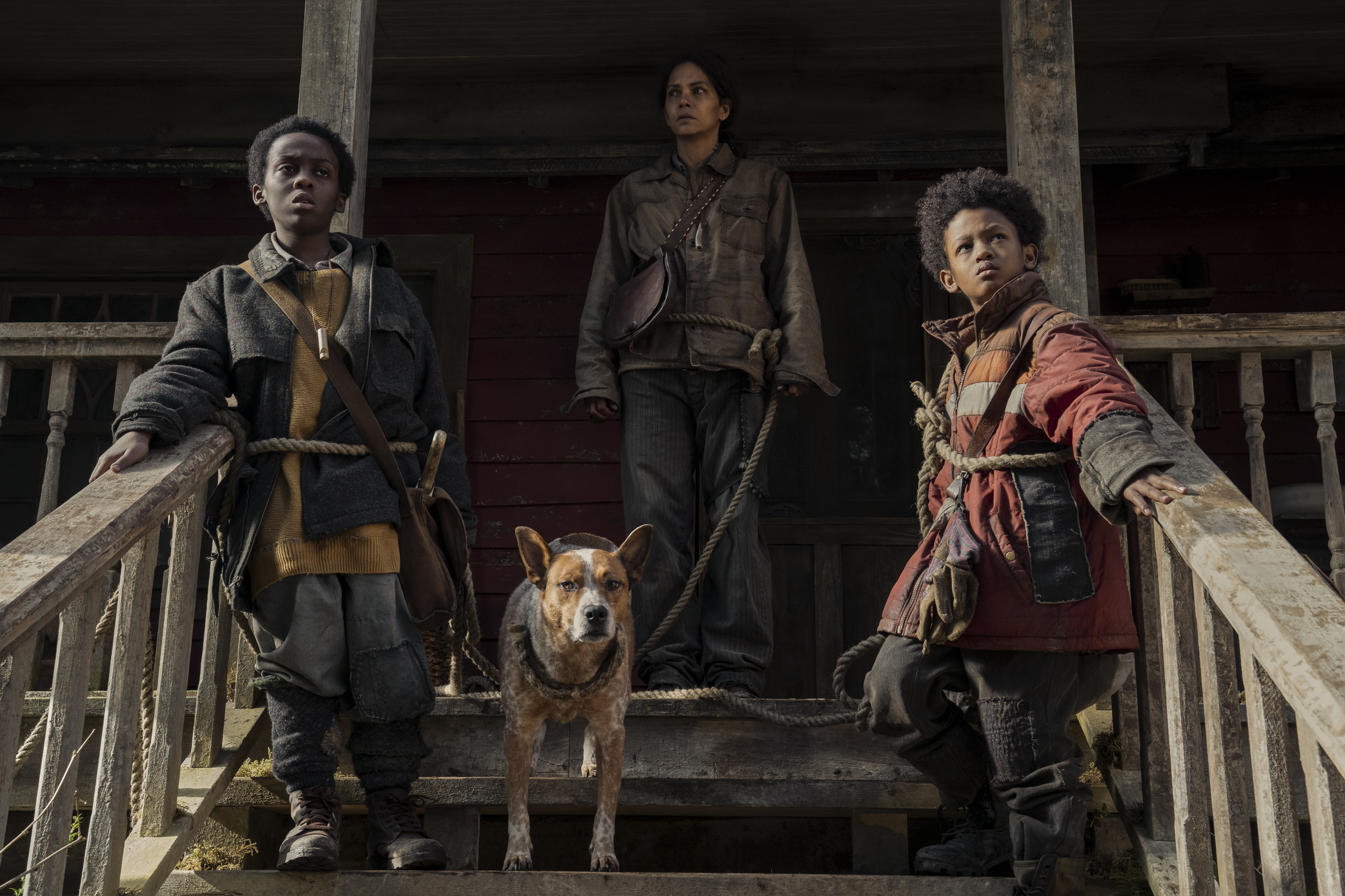 From left to right, an image of Keith L. Williams, Saniyya Sidney, Iyana Halley, and a dog standing on a porch, dressed in rugged, post-apocalyptic style clothing