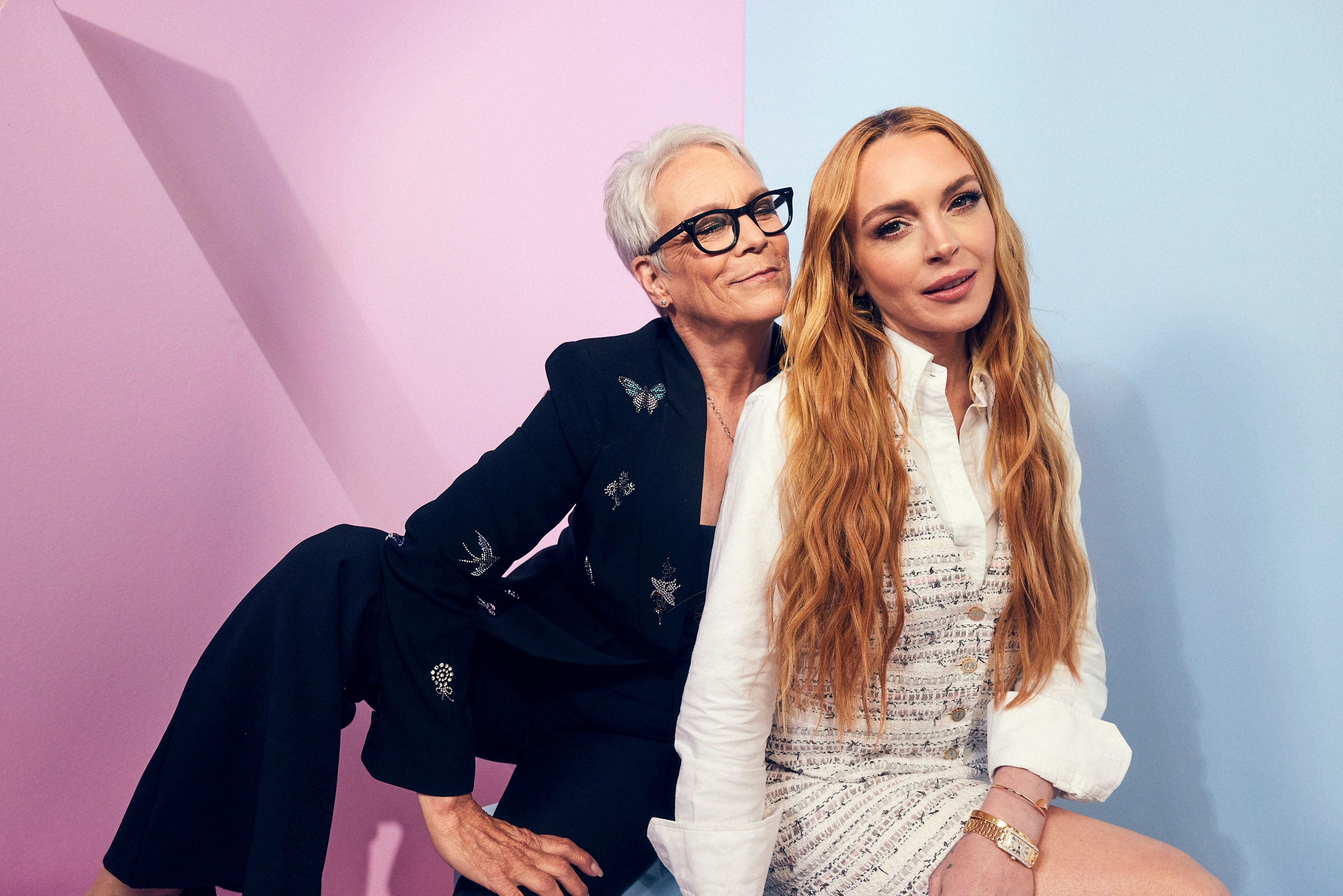 Jamie Lee Curtis in a black blazer adorned with delicate patterns poses with Lindsay Lohan in a textured white dress, both in front of pastel backdrops
