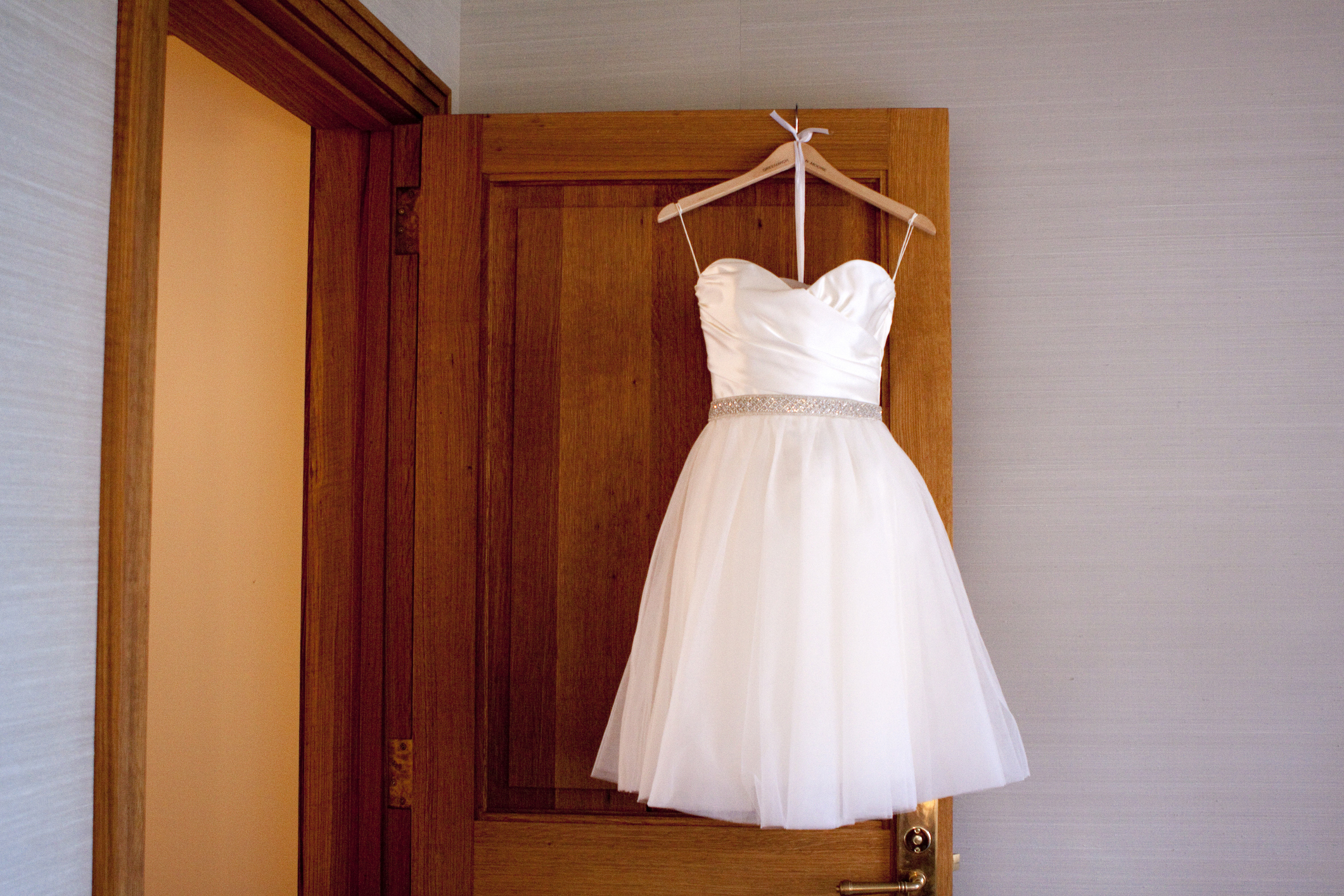 Short white dress with a strapless bodice and a silver beaded waistband, hanging on a wooden door