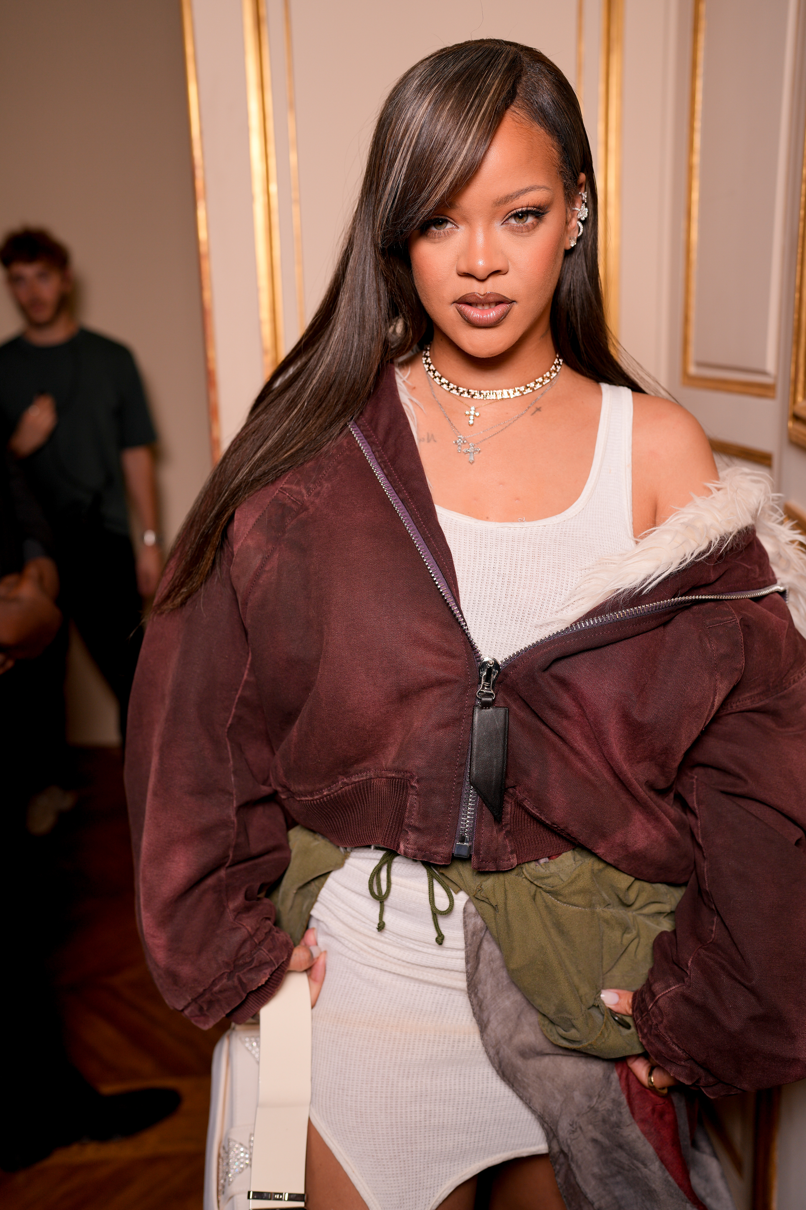 Rihanna at an event, wearing a white dress and an oversized jacket with jewelry, posing indoors