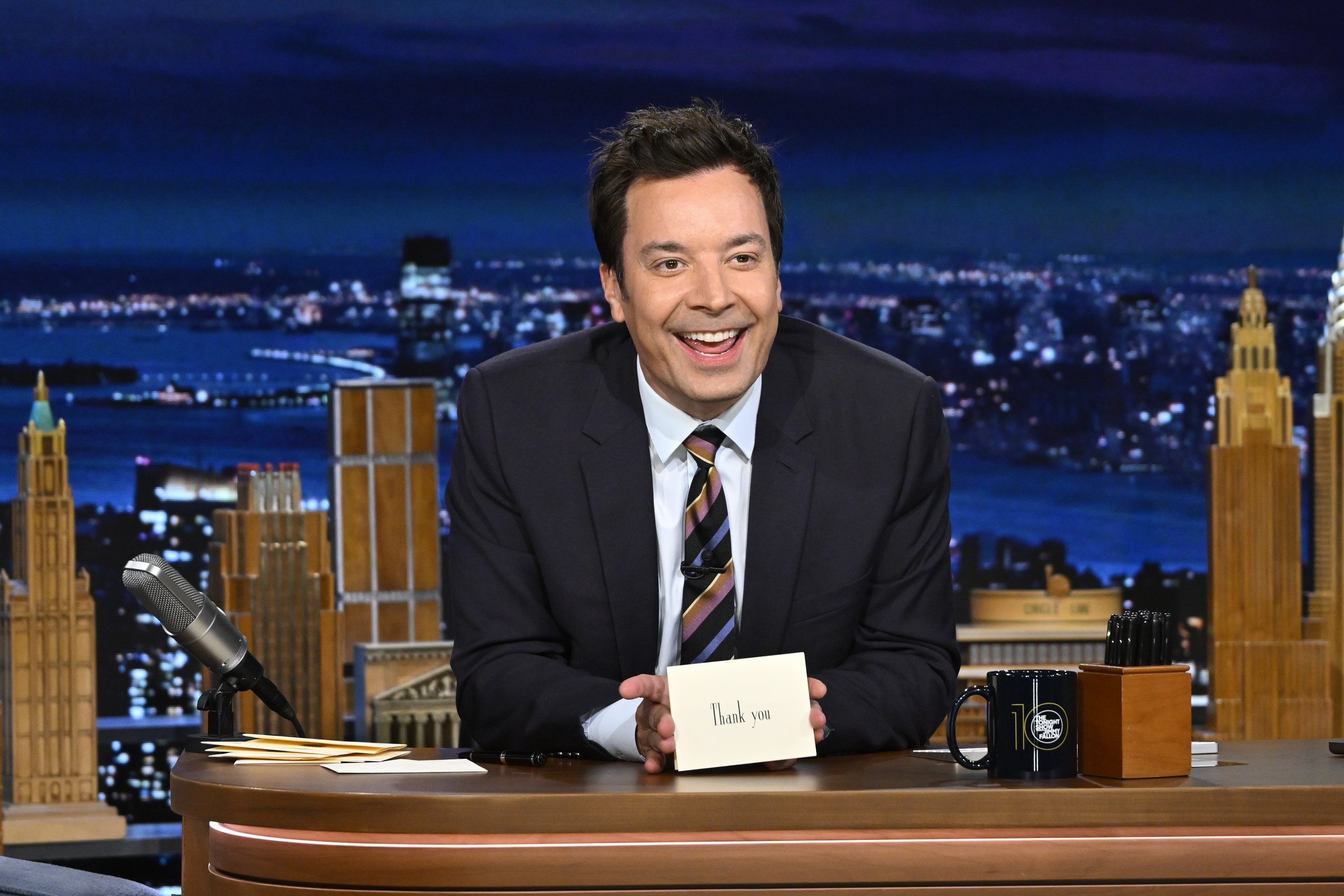 Jimmy Fallon on a late-night talk show set, smiling and holding a card with "Thank You" written on it, with a nighttime cityscape in the background