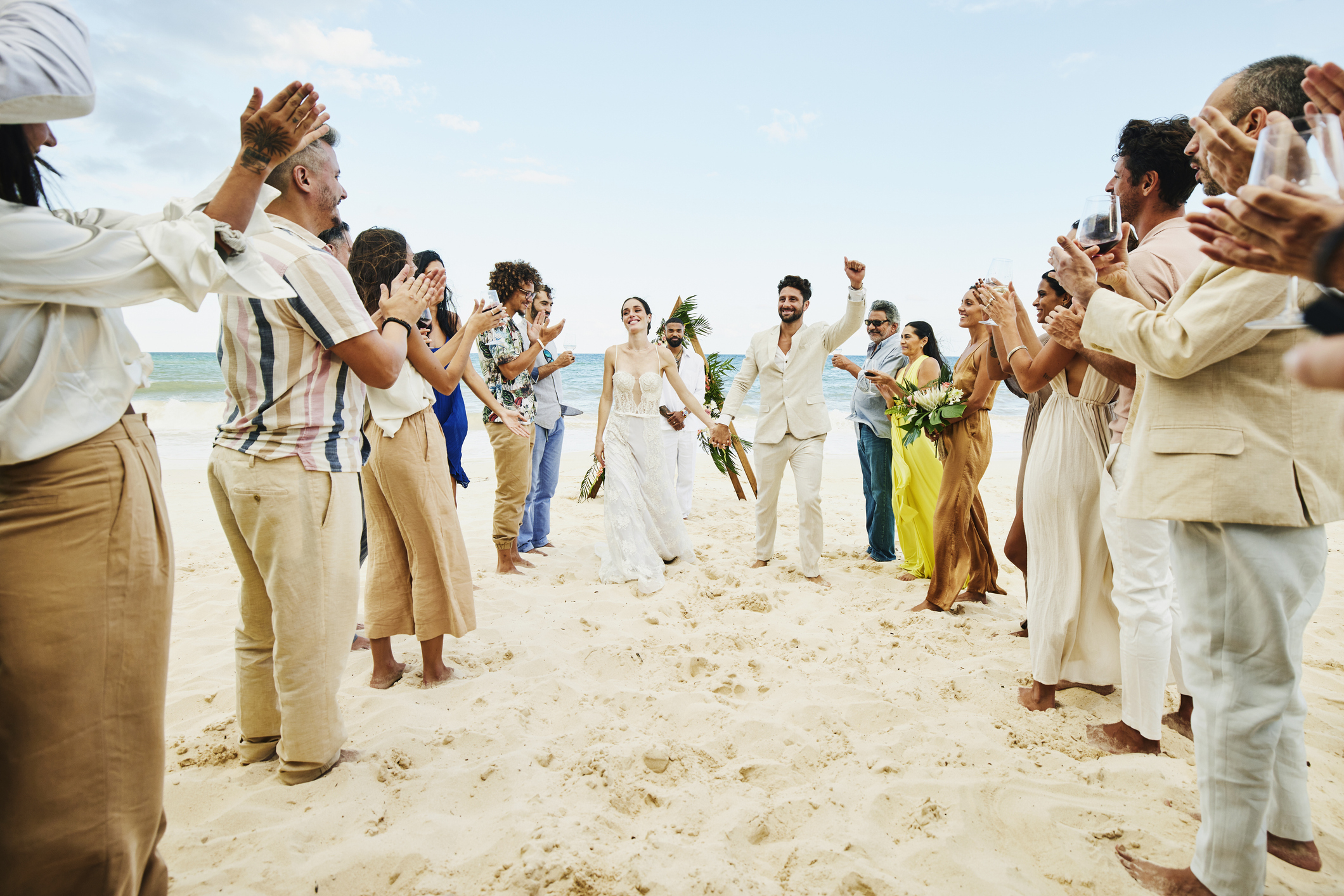 Bride and groom, both in white, walk down a sandy beach aisle surrounded by clapping guests