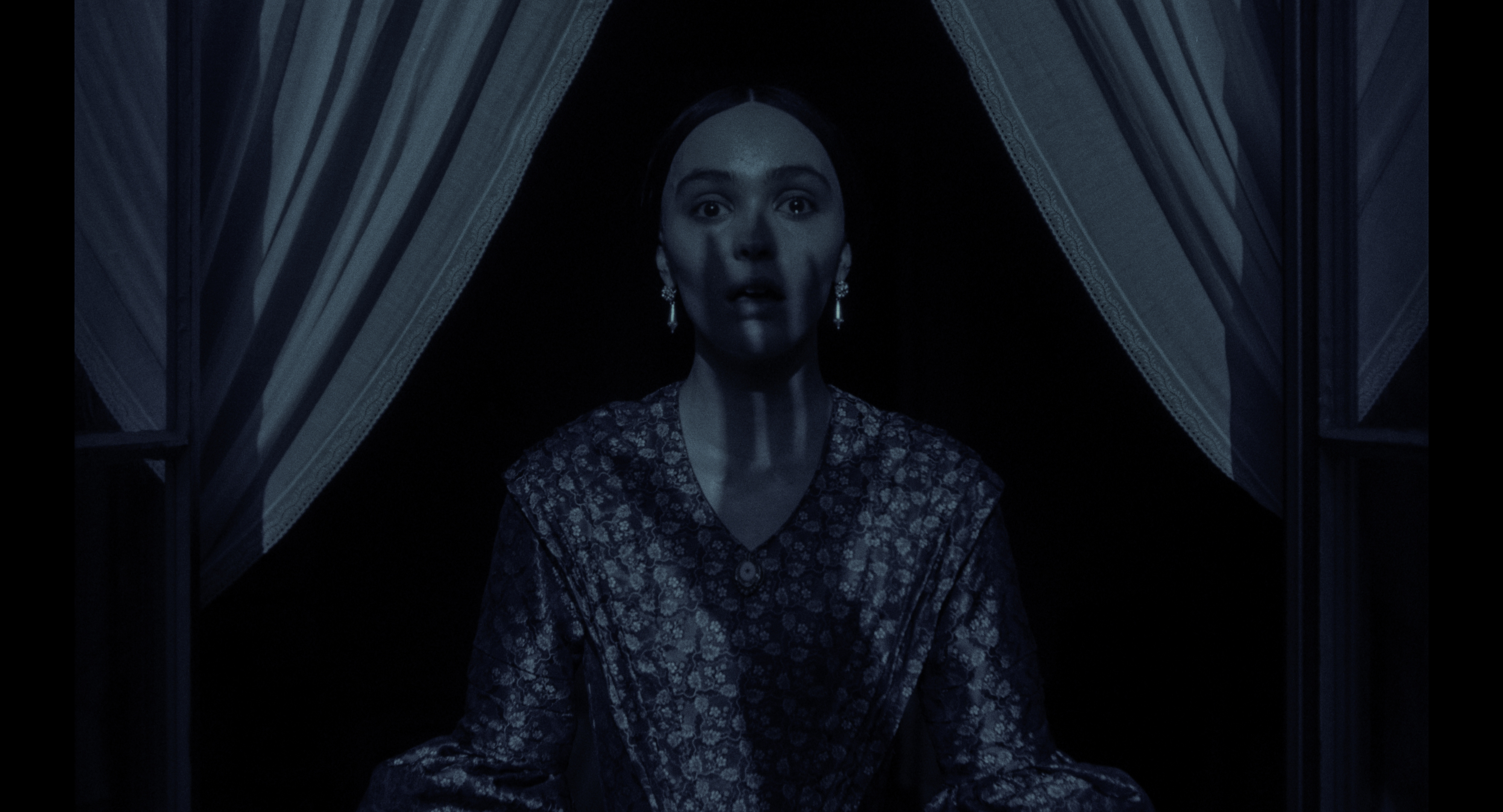 Elizabeth Olsen standing near a window, wearing a patterned robe, in a scene with dramatic lighting