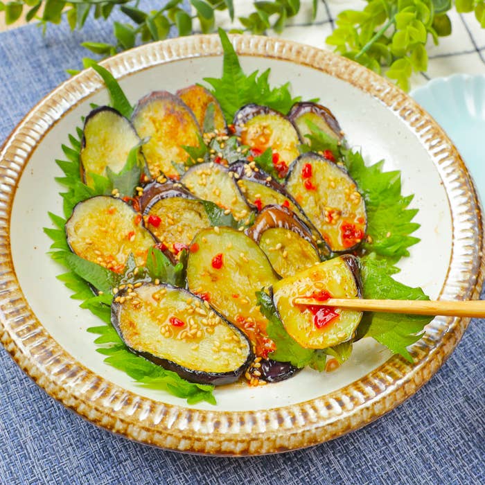 皿に盛られたナスの料理が、新鮮なシソの葉の上に載せられ、ゴマと唐辛子が散りばめられた様子が写っています。