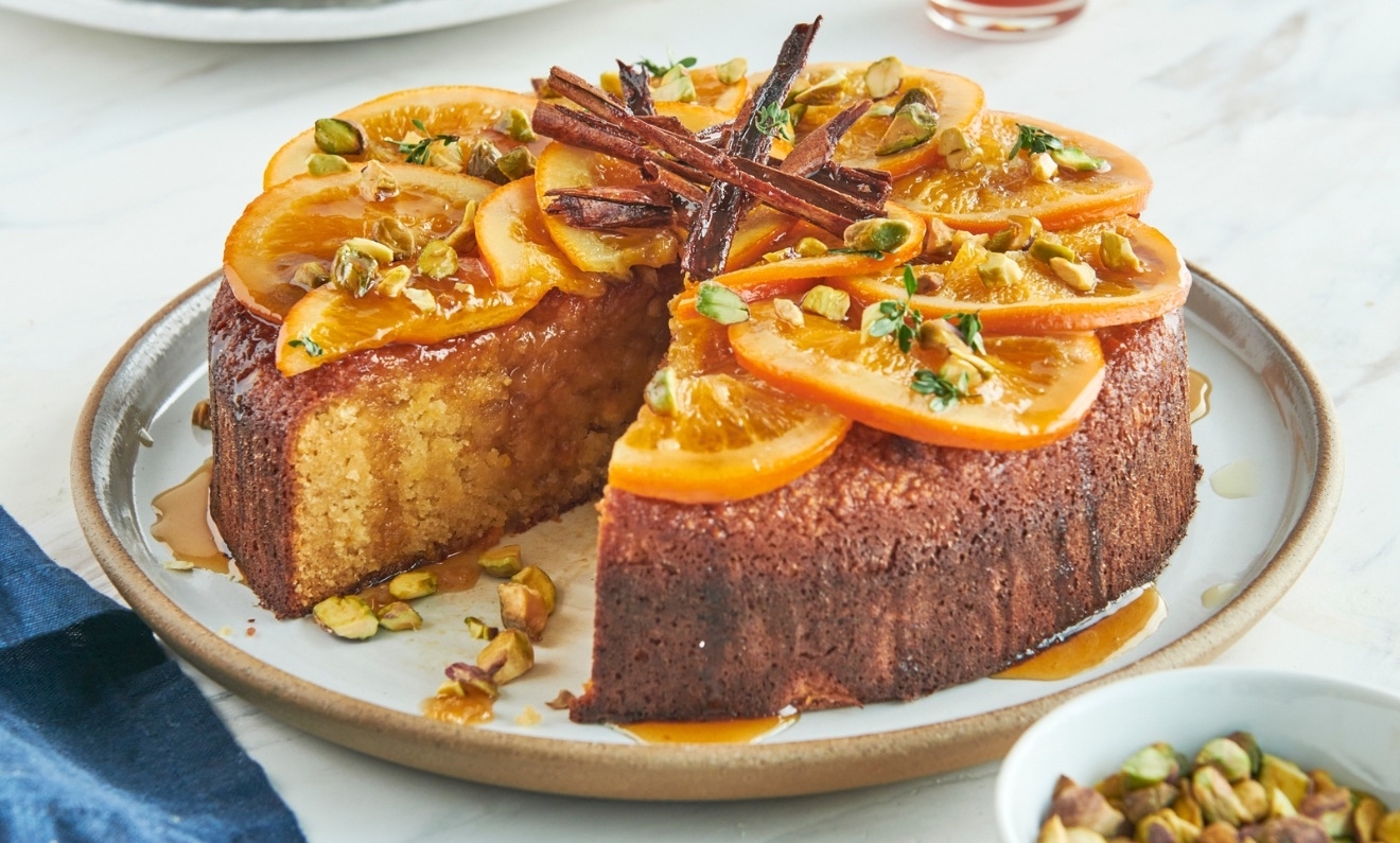A round orange-pistachio cake topped with caramelized orange slices, pistachios, and cinnamon sticks, with a slice cut out, displayed on a white plate